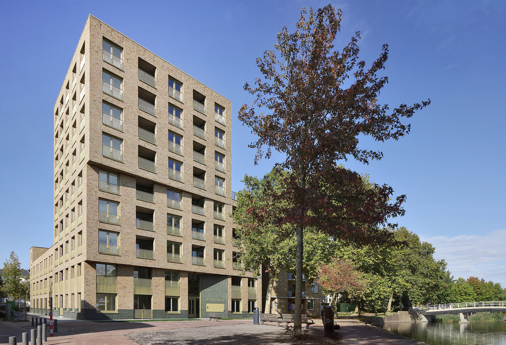 Tower volume in buff brick with deep balconies beside an autumn tree and canal edge