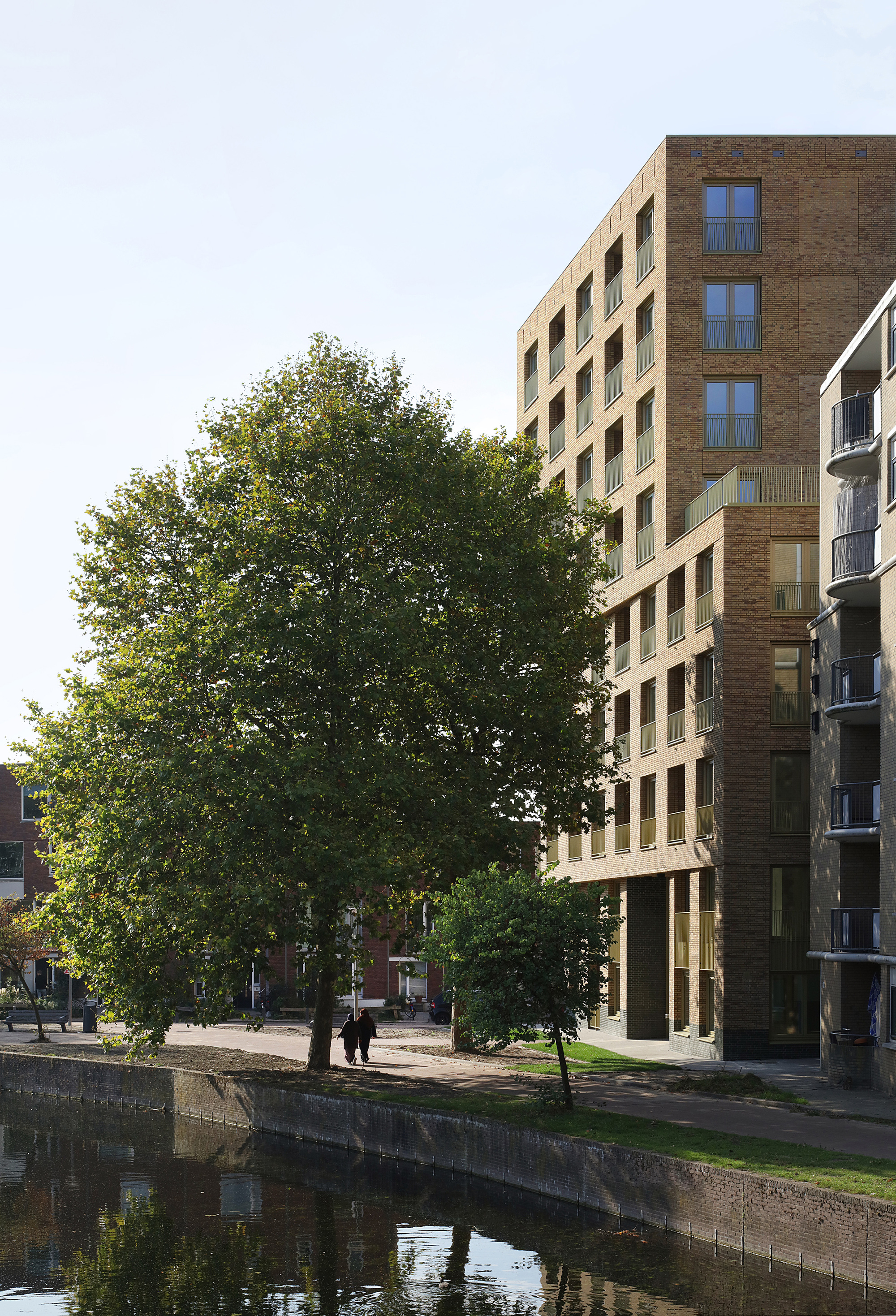 Brick facade with recessed balconies rising behind a canal lined with mature trees