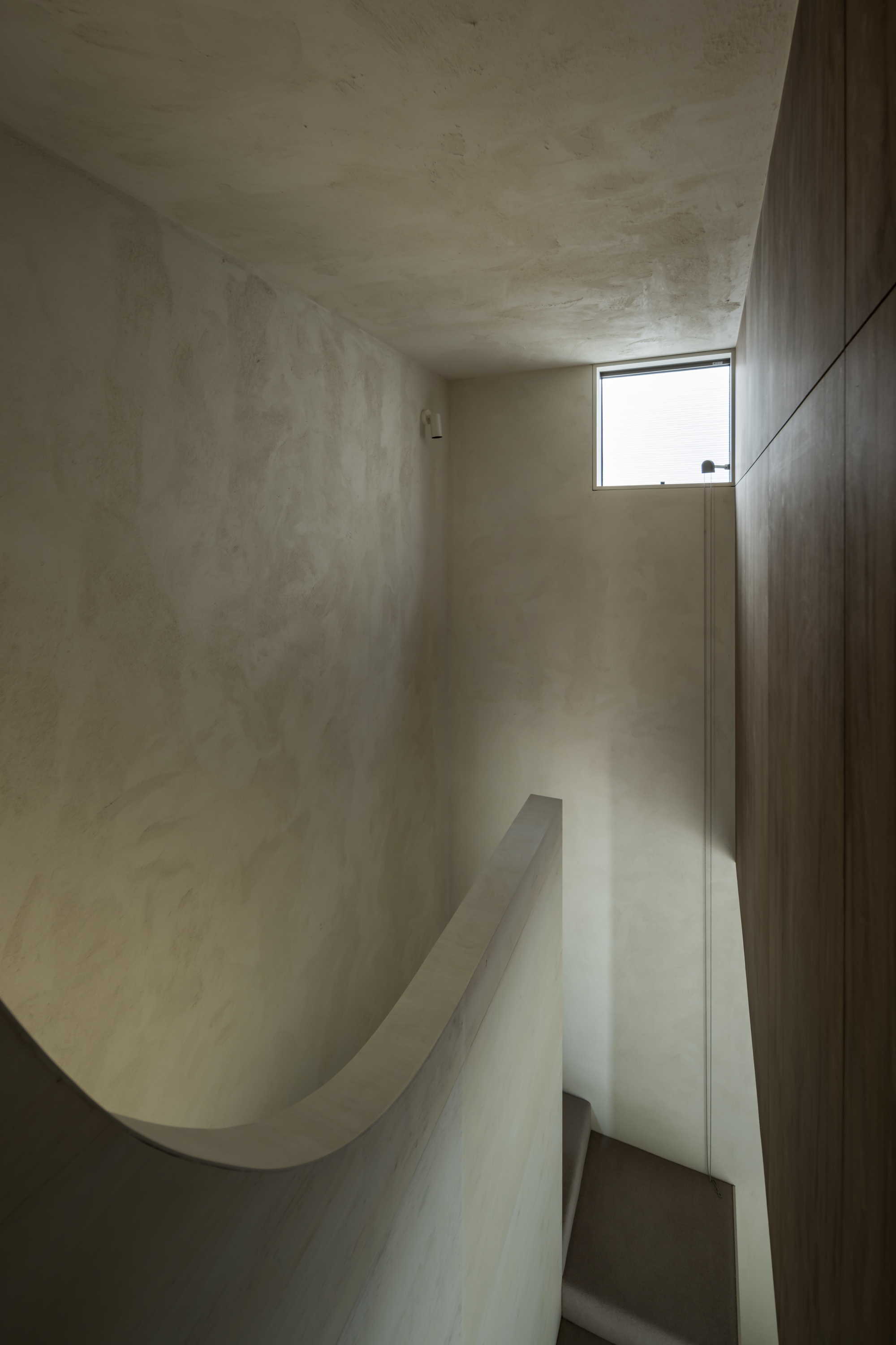 Curved plaster stairwell with dark timber wall and narrow horizontal window casting daylight