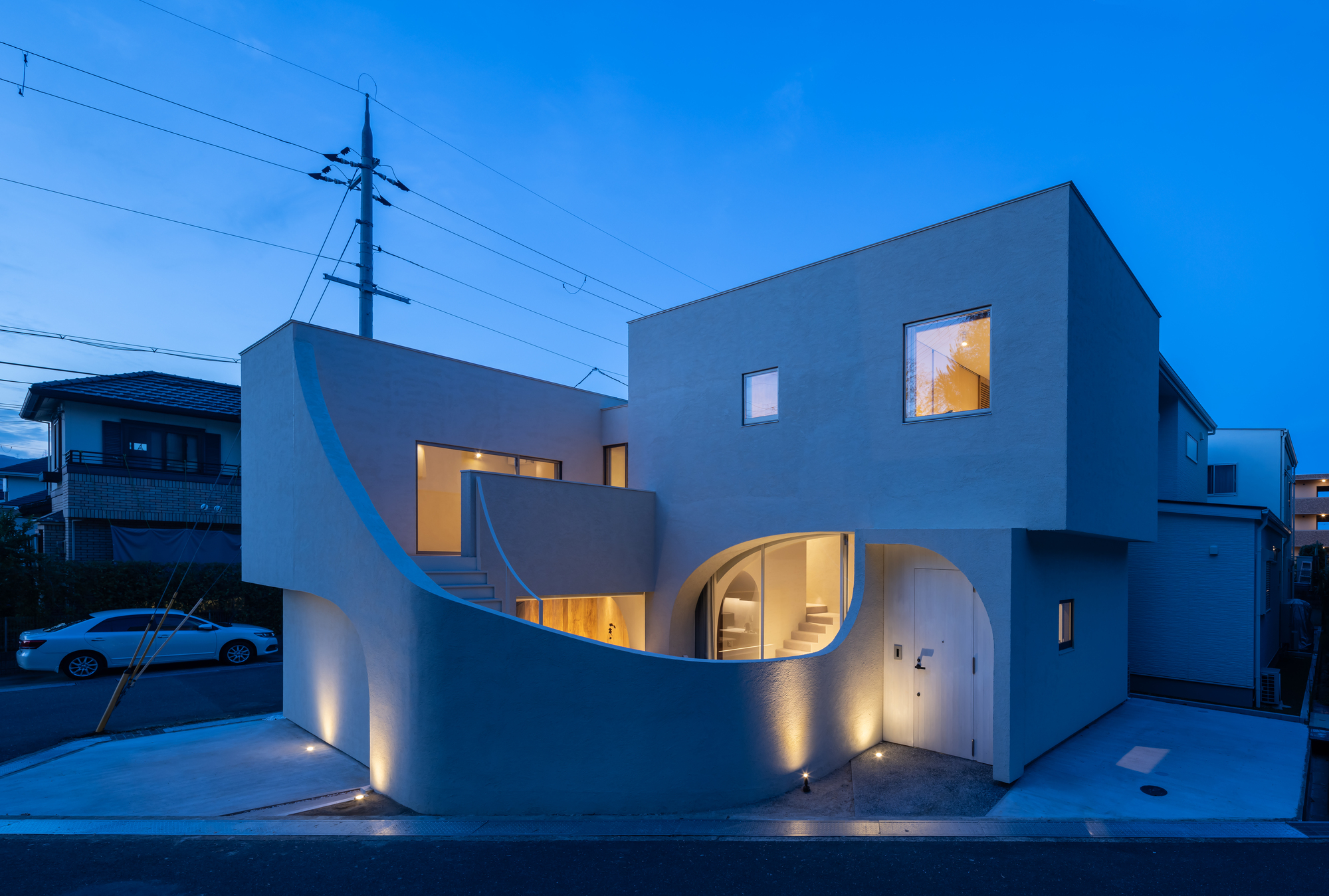 Street view of the grey facade with curved openings and uplighting at blue hour