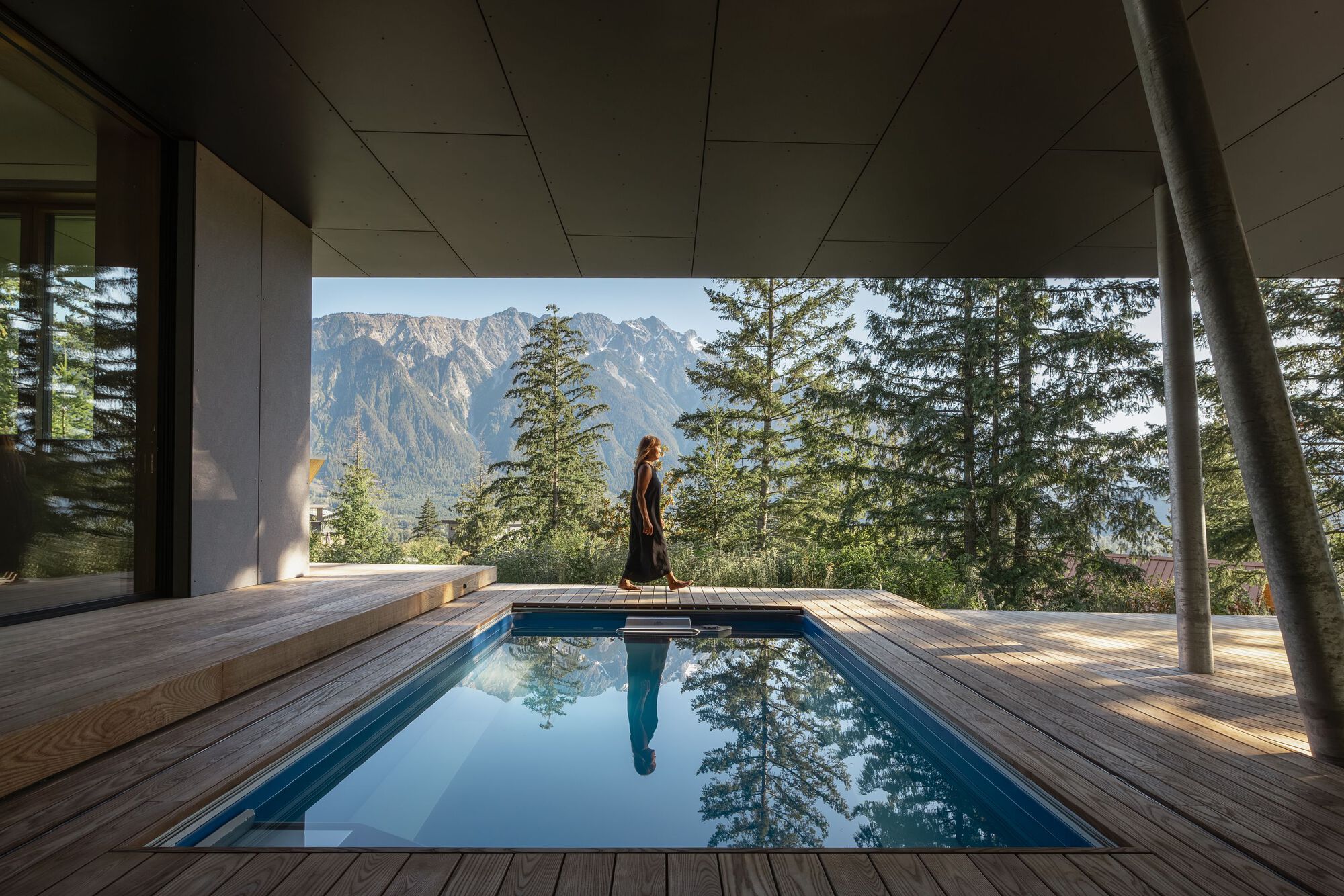 Lap pool on timber deck framing mountain views through slender steel columns and evergreen trees