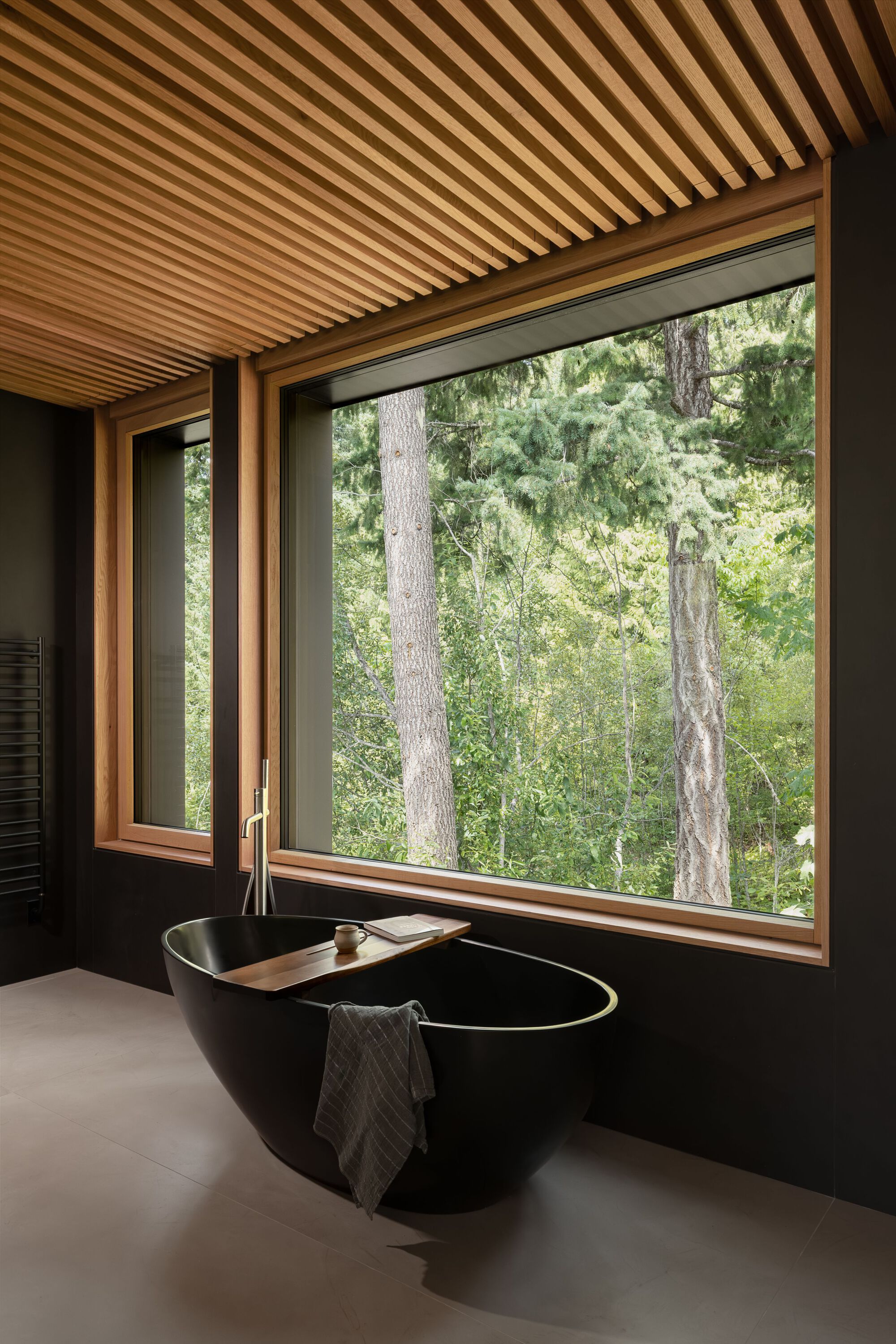 Freestanding black bathtub beneath a slatted timber ceiling and wood-framed window with forest view