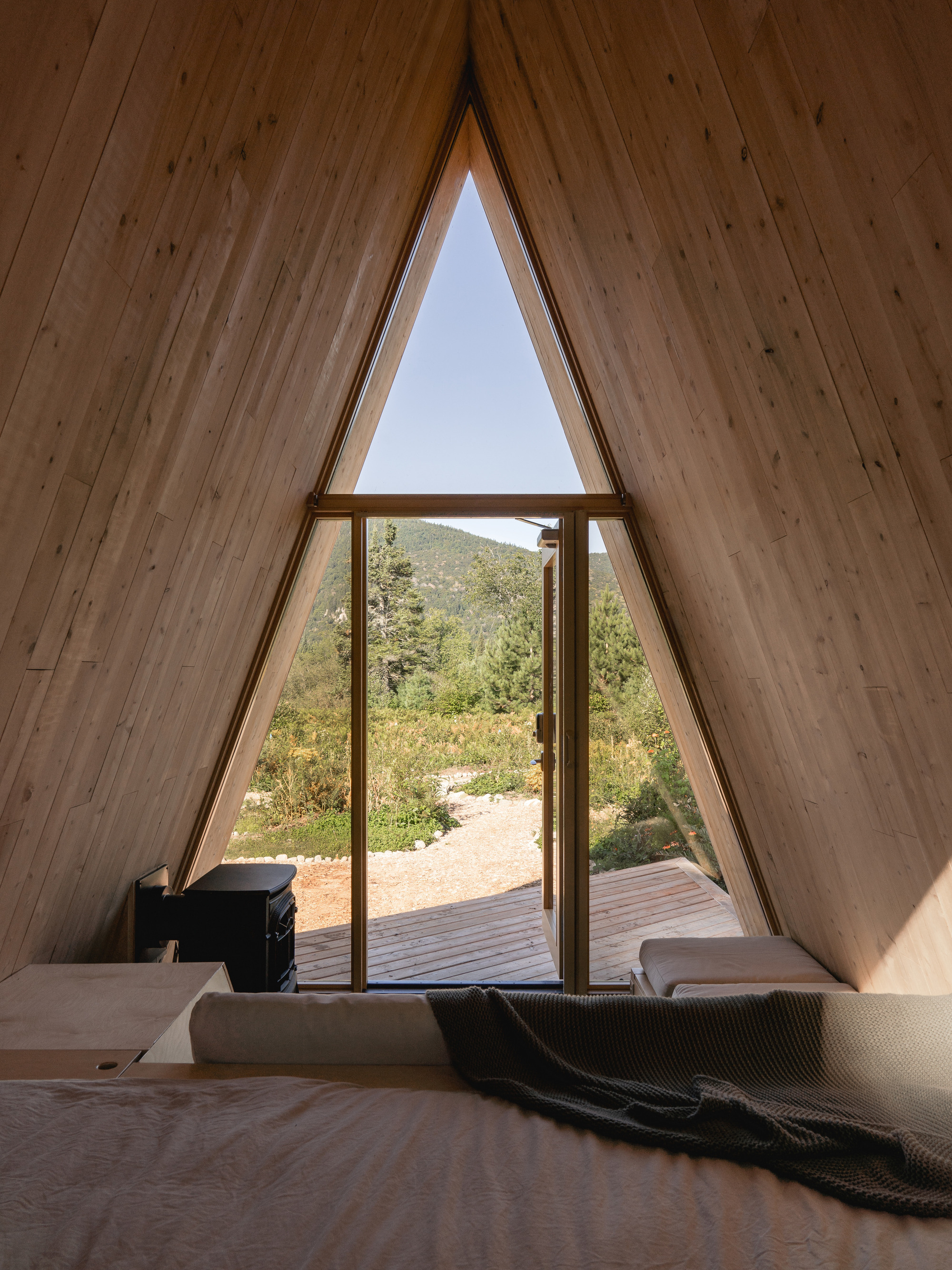 Interior view through triangular glazed opening framing timber deck and mountain landscape beyond a bed
