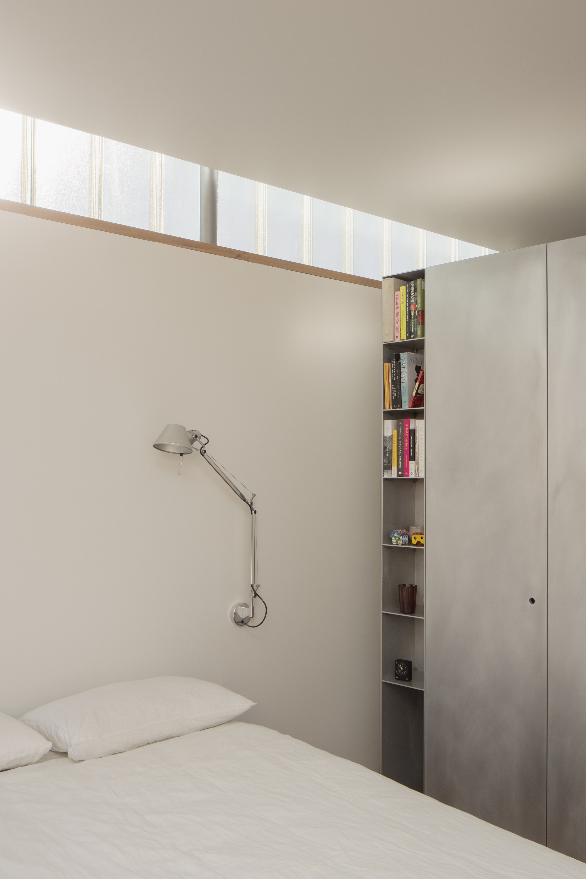 Bedroom with clerestory window above bed and narrow vertical bookshelf beside plywood cabinetry