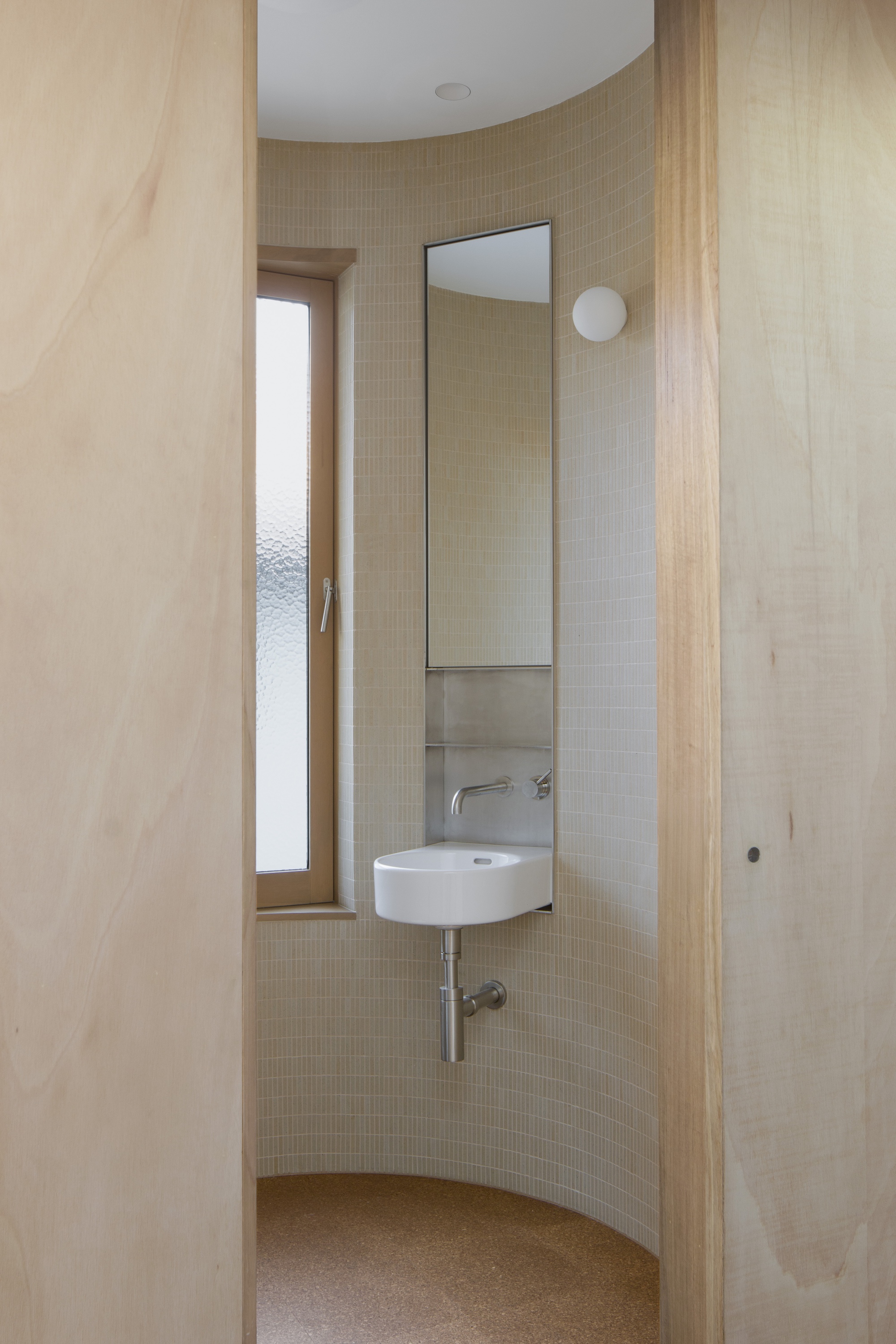 Curved powder room with pale mosaic tile walls and a wall-mounted basin beneath a vertical mirror