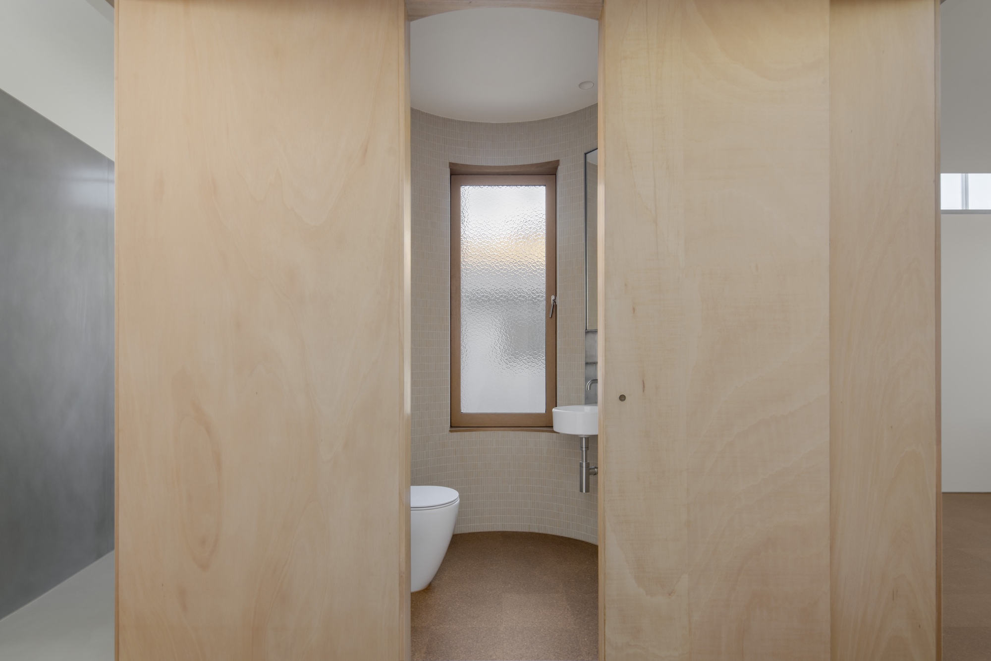 Curved bathroom wall clad in light plywood panels with translucent glass door at center