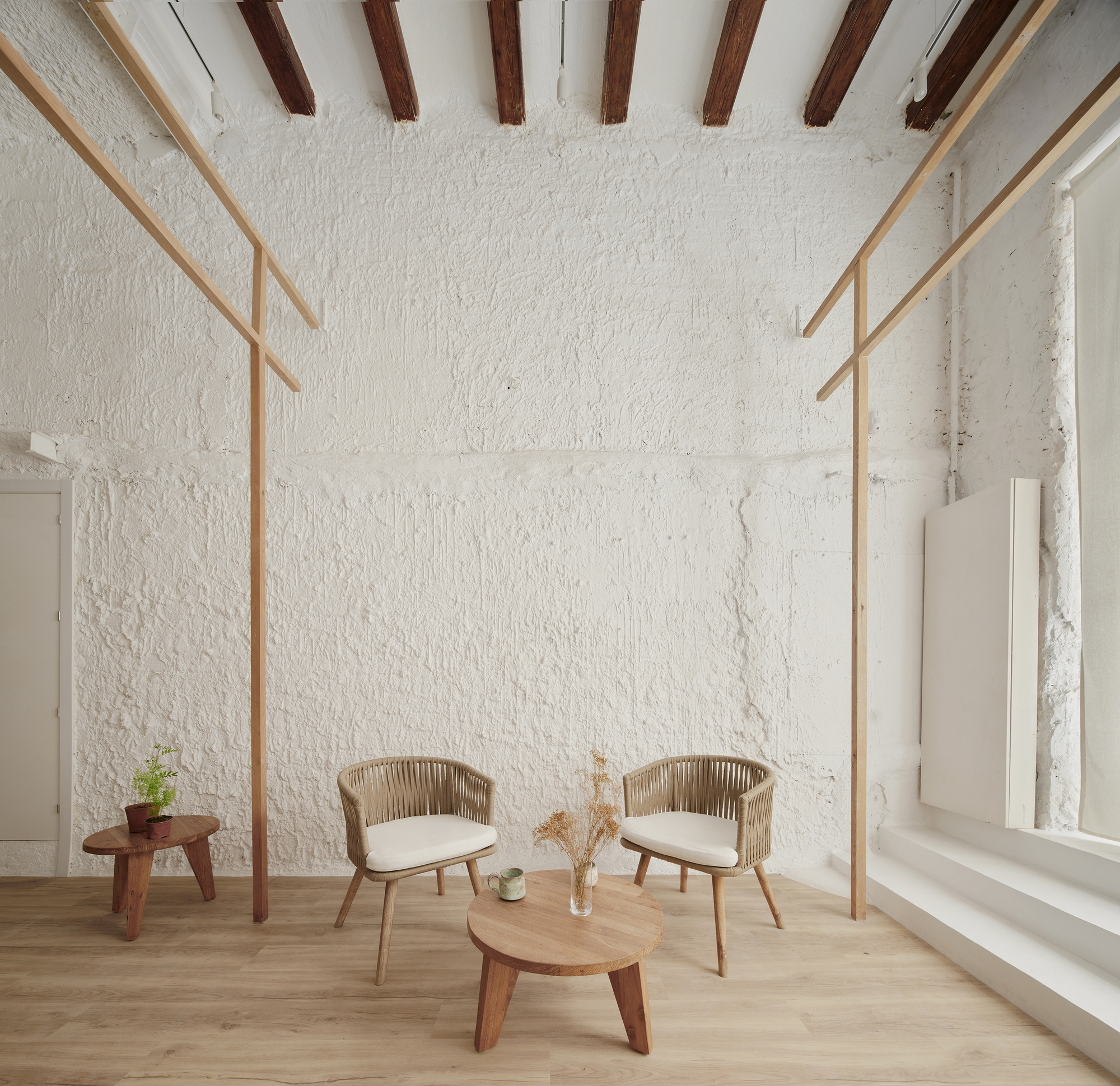 Seating area beneath exposed timber ceiling beams with textured white plaster walls and a tall window