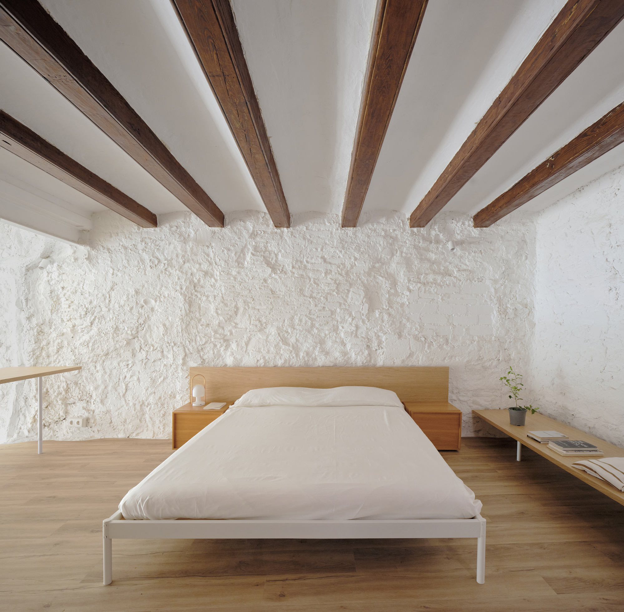 Bedroom with exposed timber ceiling joists above a timber platform bed against a rough white plaster wall