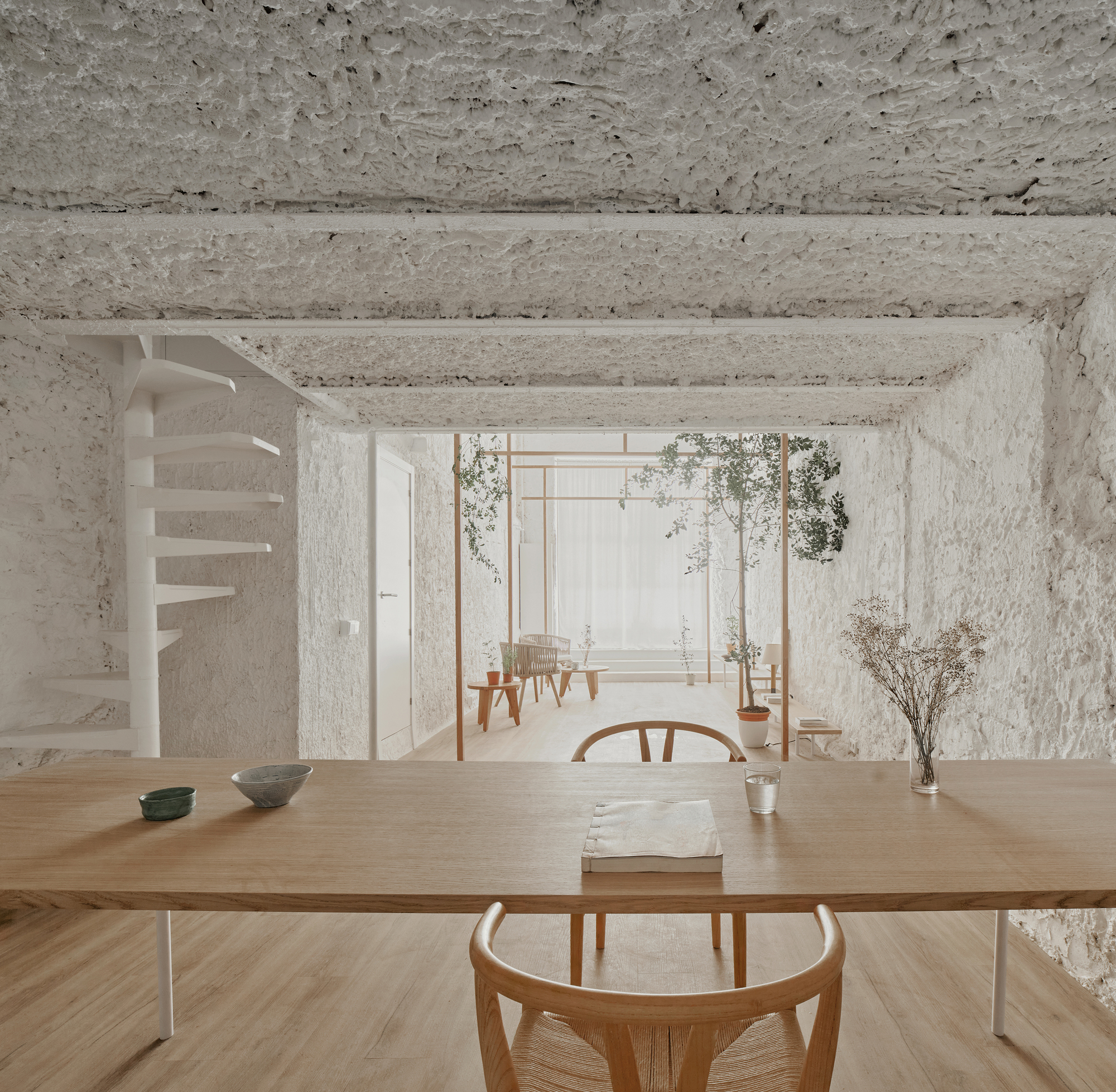 View across dining table toward spiral stair and timber pergola with potted plants in soft daylight