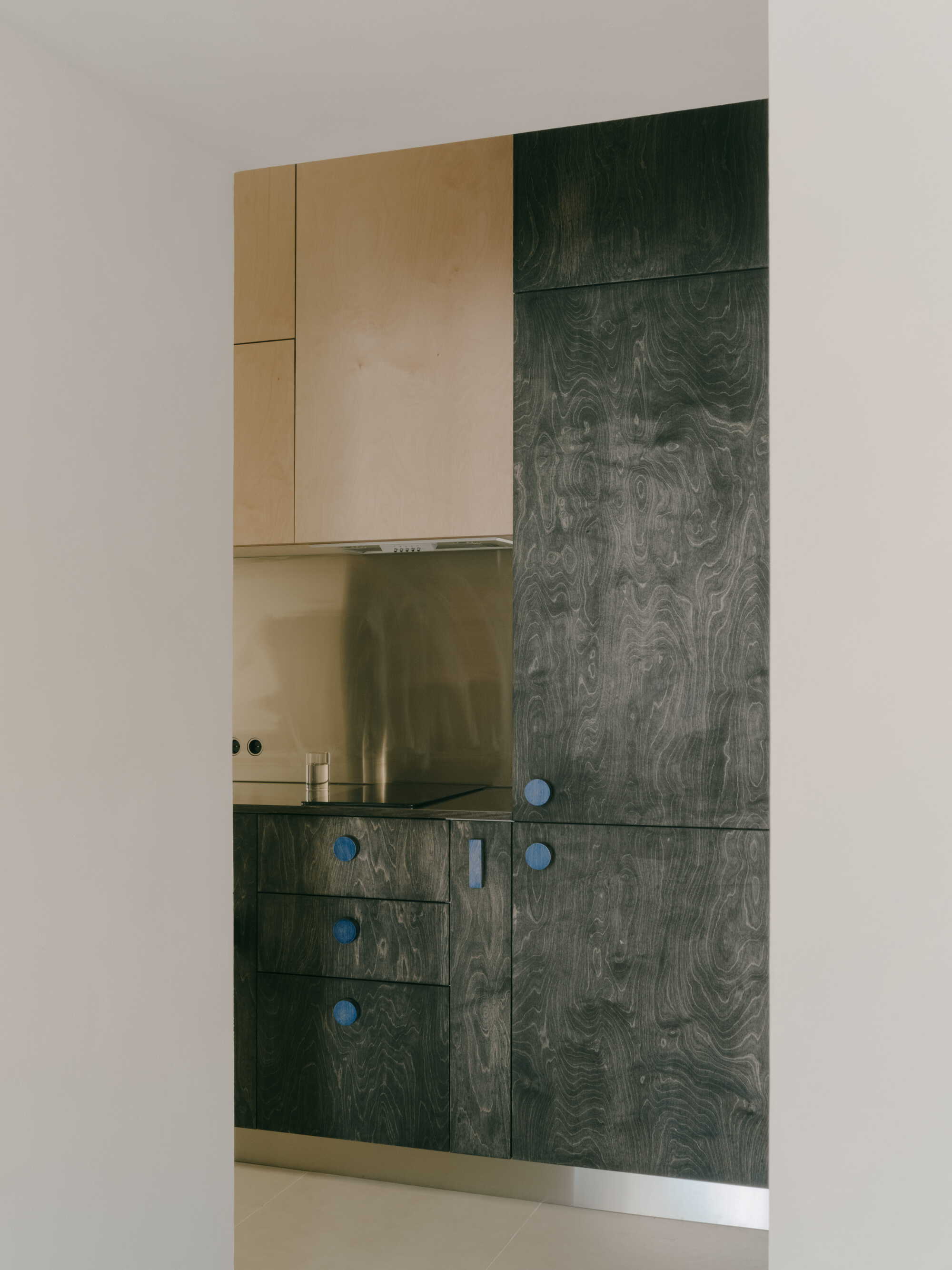 Kitchen cabinetry with dark wood veneer doors, blue knobs, and reflective backsplash under light wood upper units