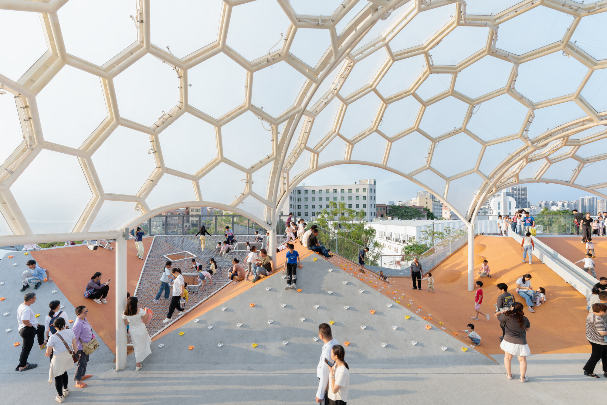 Interior view beneath the hexagonal dome canopy with visitors gathering on orange play surfaces and planted areas