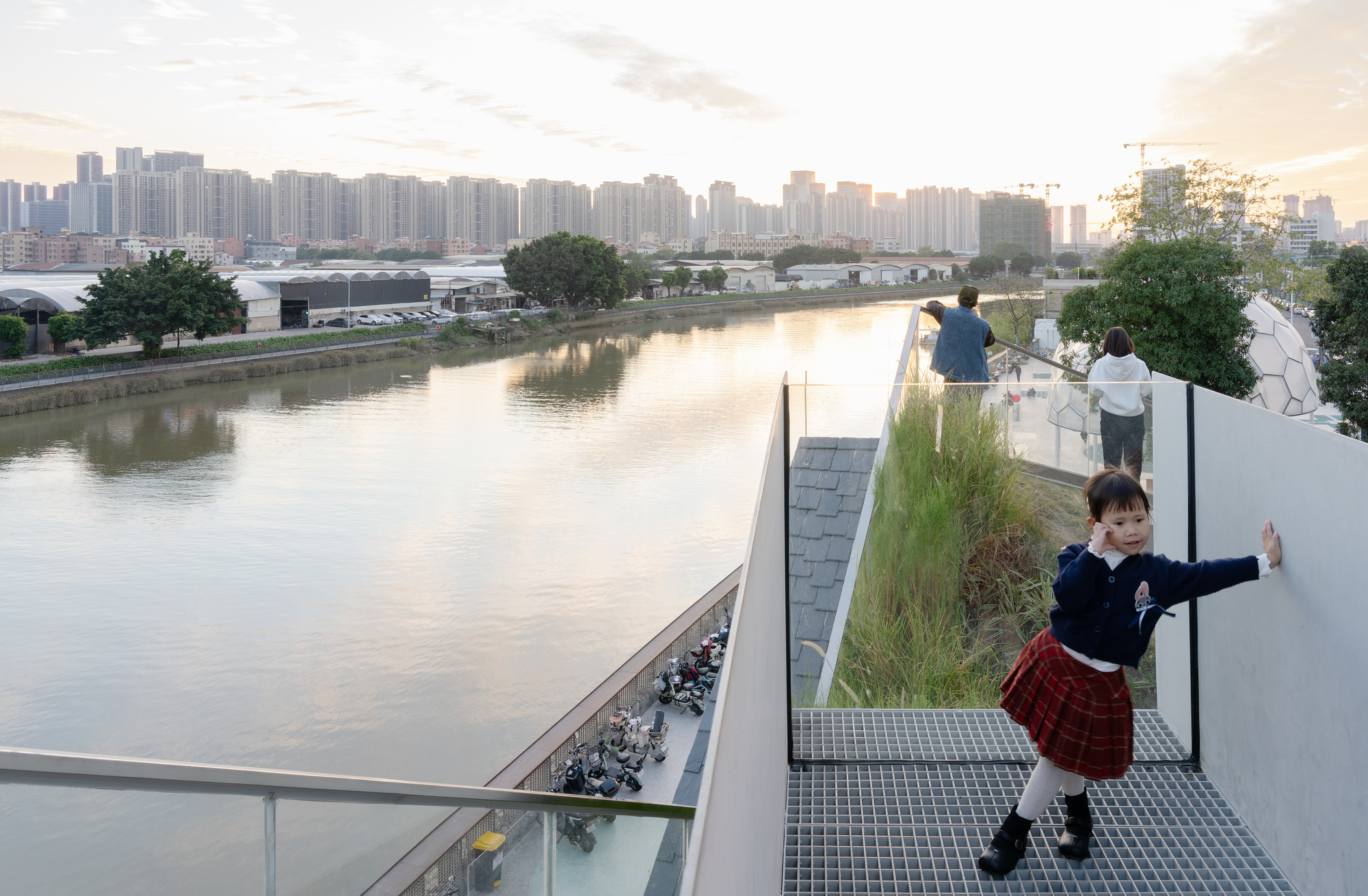 Elevated walkway with planted grasses overlooking the canal at sunset with visitors along the glass balustrade