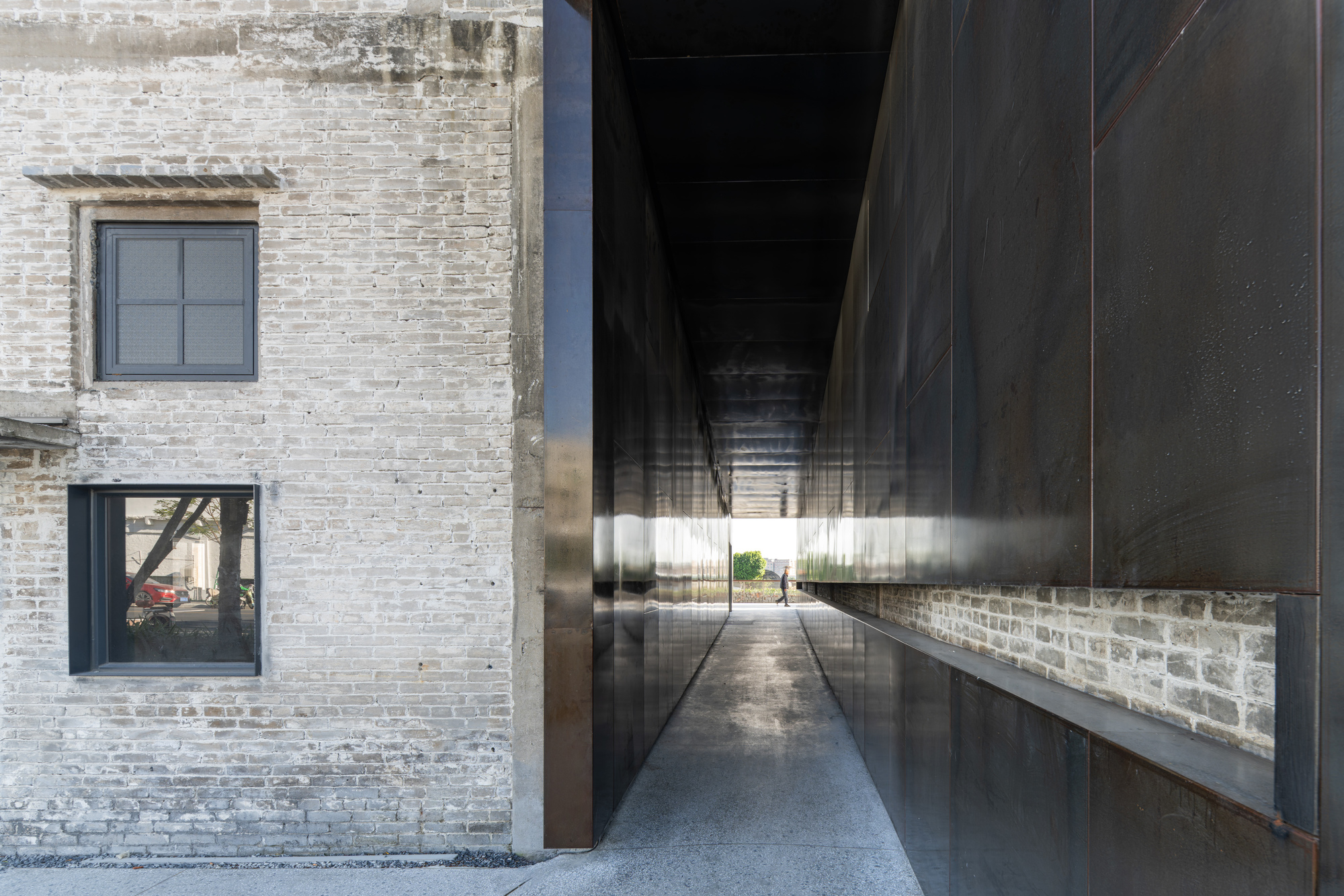 Passageway between weathered brick wall and blackened steel panel reflecting the opening ahead