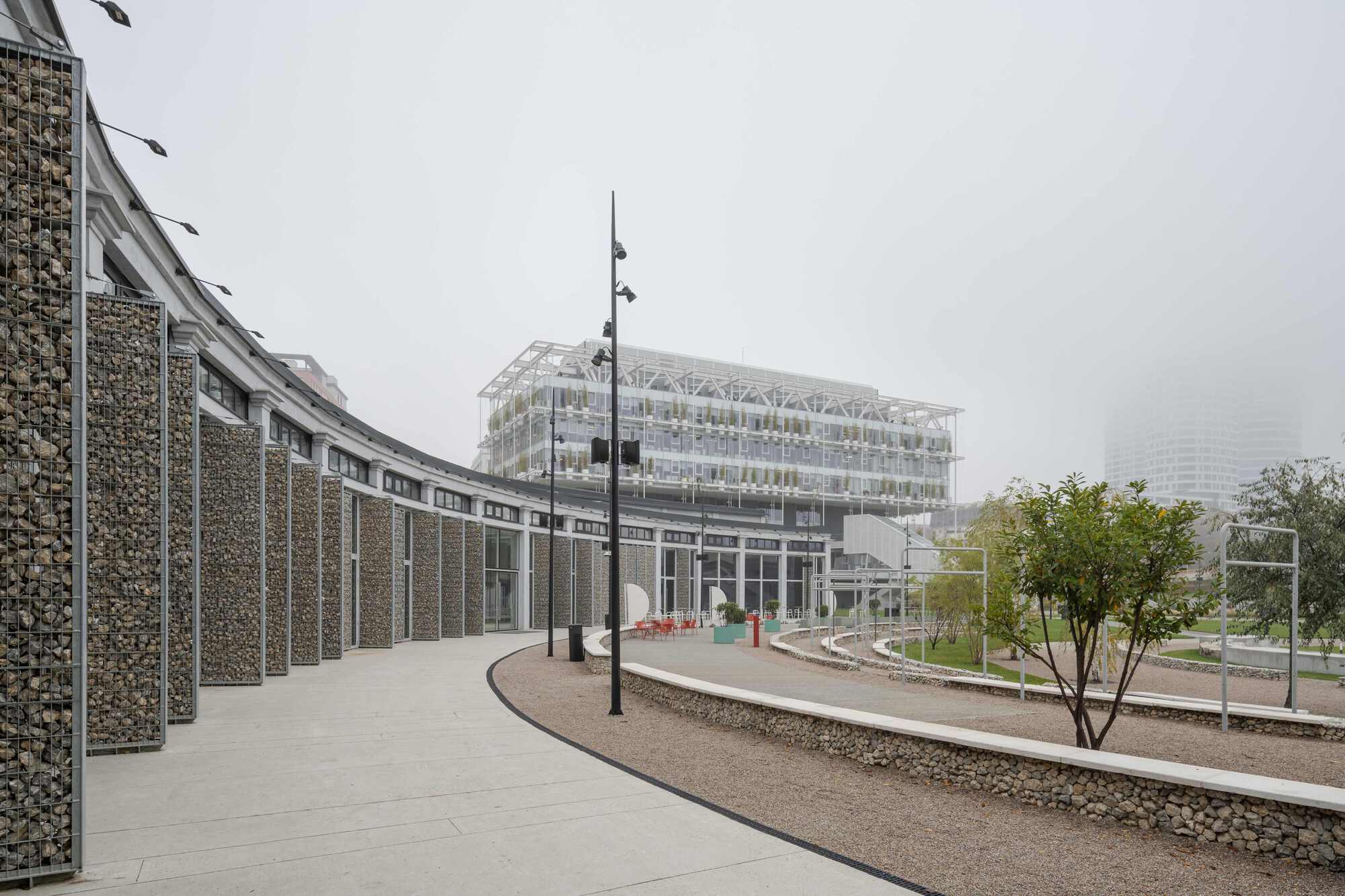 Curving path alongside a gabion wall leading toward the greenhouse under overcast sky