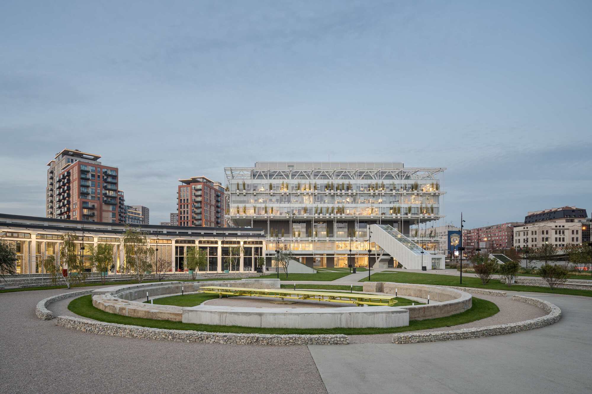 Circular sunken plaza with grass and gravel rings in front of the transparent greenhouse building at dusk