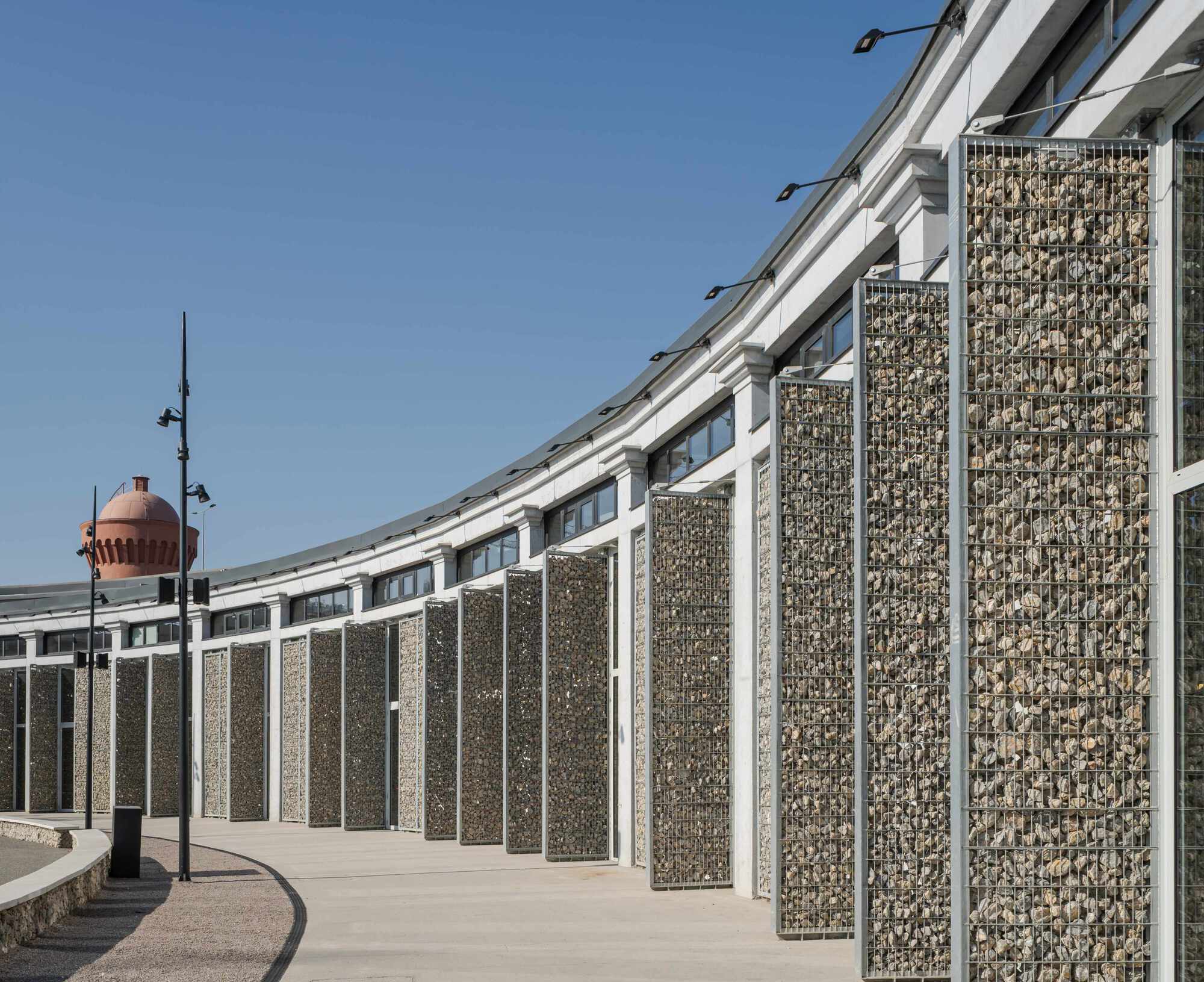 Curved gabion wall filled with stone set in steel frames along an exterior walkway under blue sky