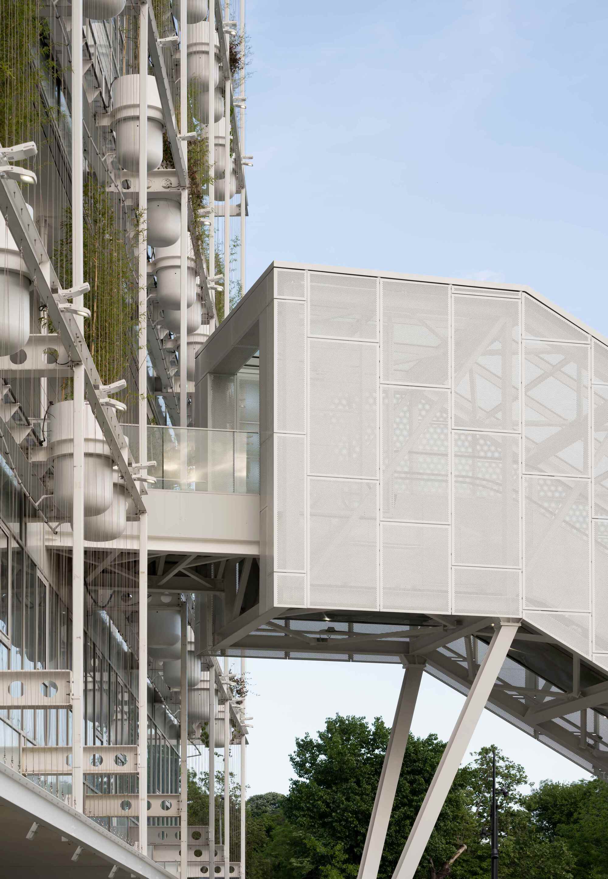 Cantilevered volume with translucent paneled facade supported by diagonal white steel trusses above green trees