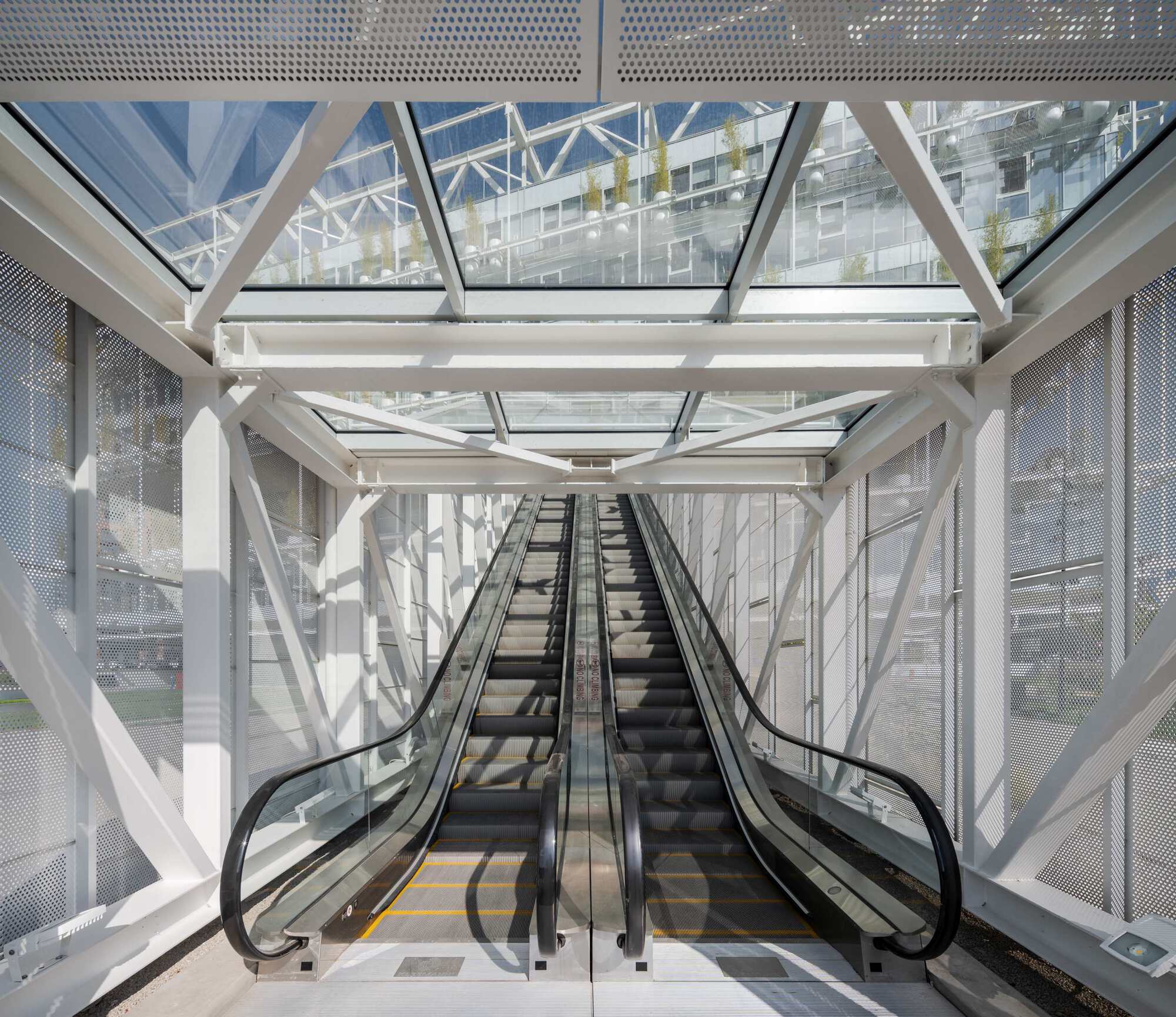 Escalator shaft enclosed by white steel trusses and perforated metal screens with views to courtyard beyond