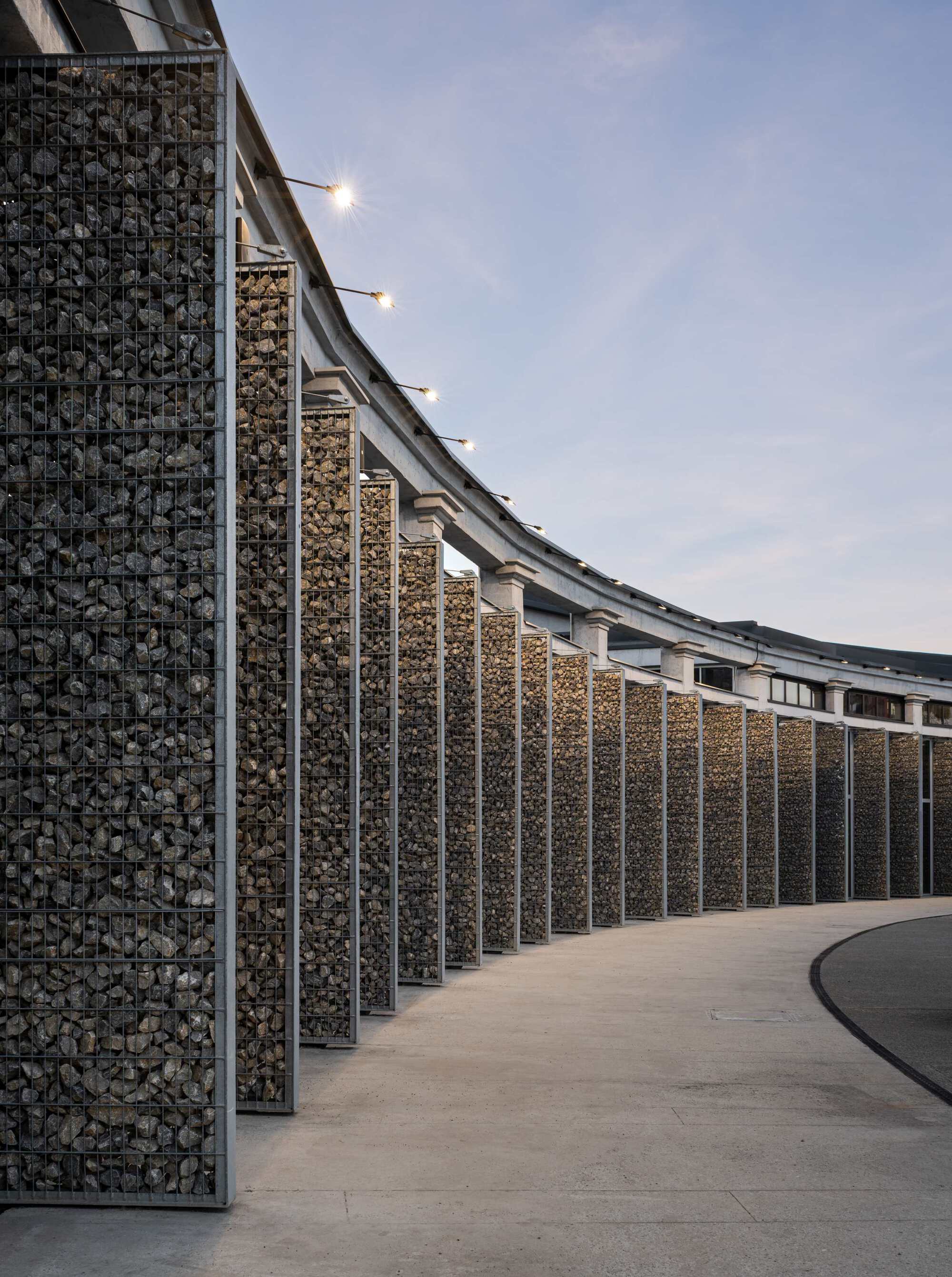 Row of gabion columns curving along a walkway beneath a highway deck at twilight