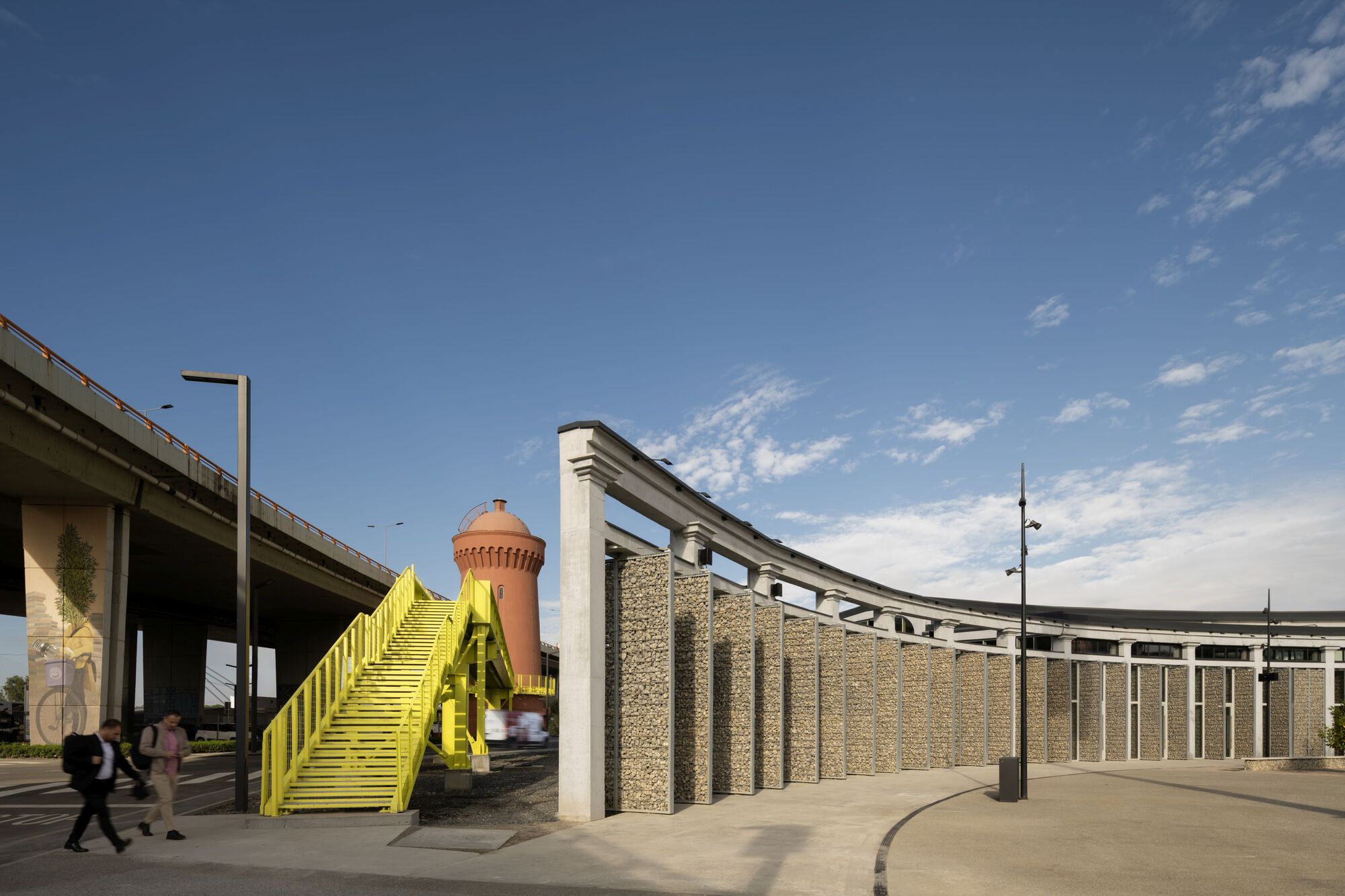 Yellow metal staircase beside a gabion wall and pink water tower under a concrete overpass
