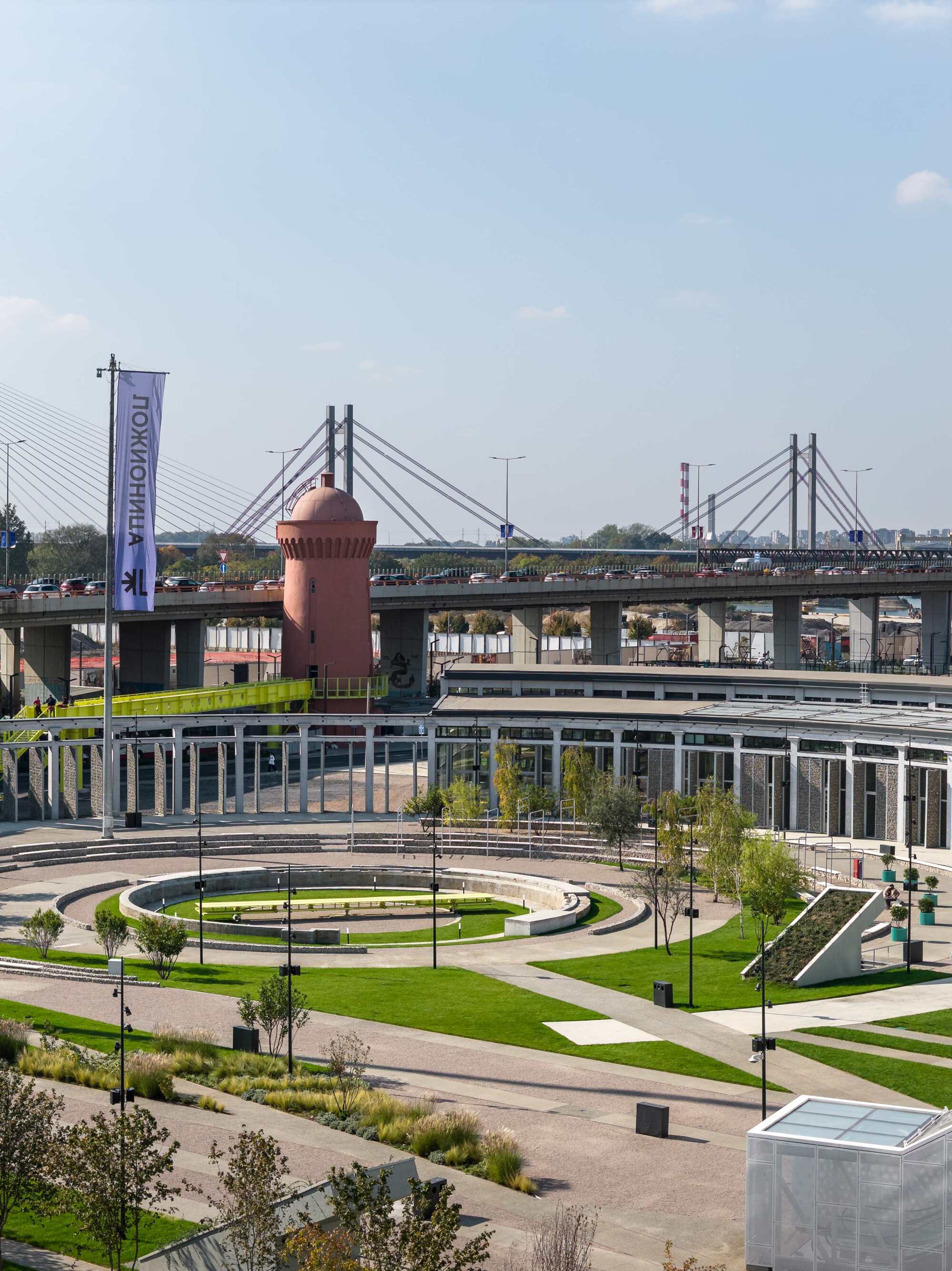 Circular landscaped plaza with lawn rings and pink water tower beneath cable-stayed bridges