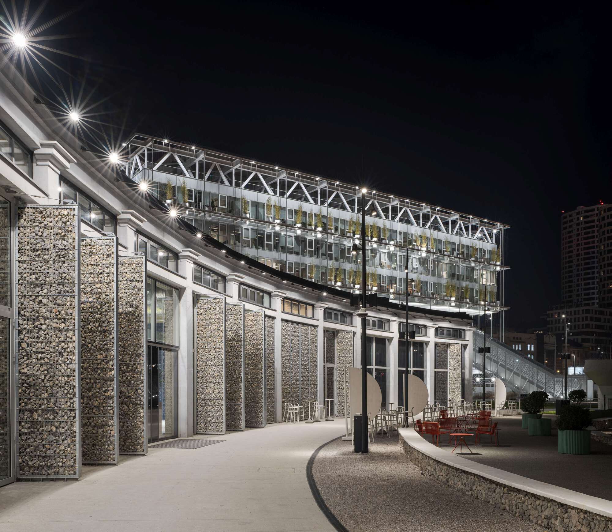 Gabion-filled columns and glass pavilion beneath an illuminated highway viaduct at night