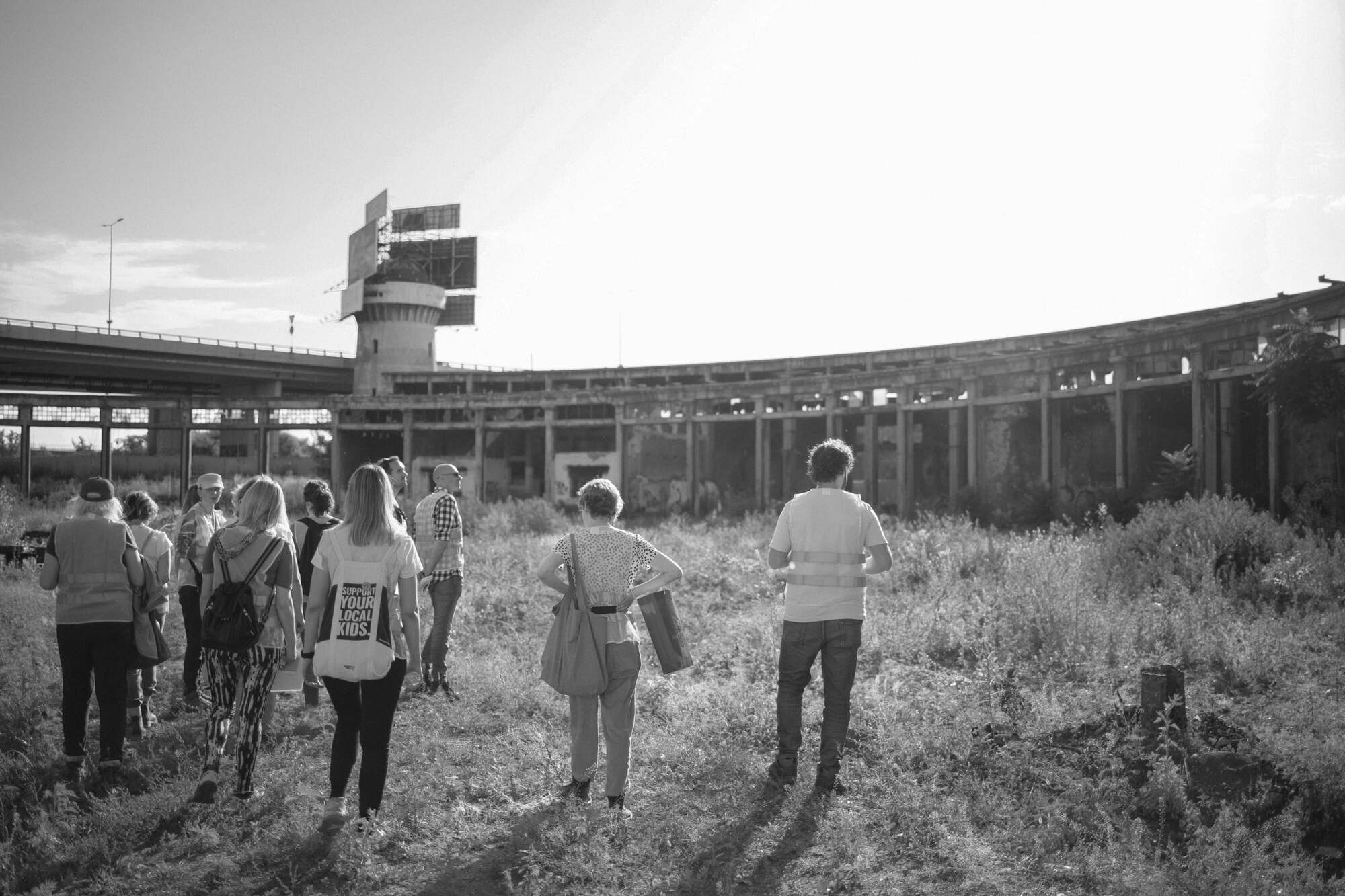 Group of visitors walking through overgrown grasses towards a curved colonnade and control tower at dusk