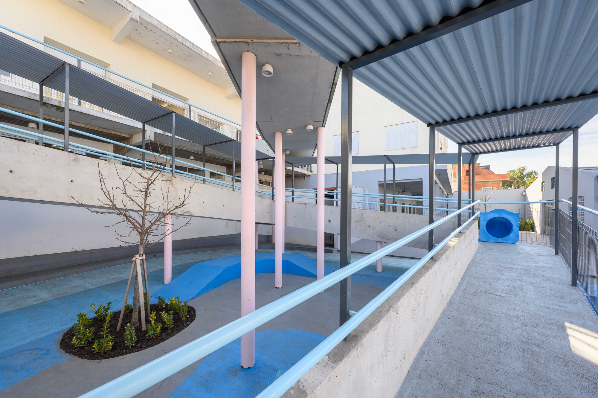 Concrete ramp with pink cylindrical columns and corrugated metal canopy shading a planted circular bed