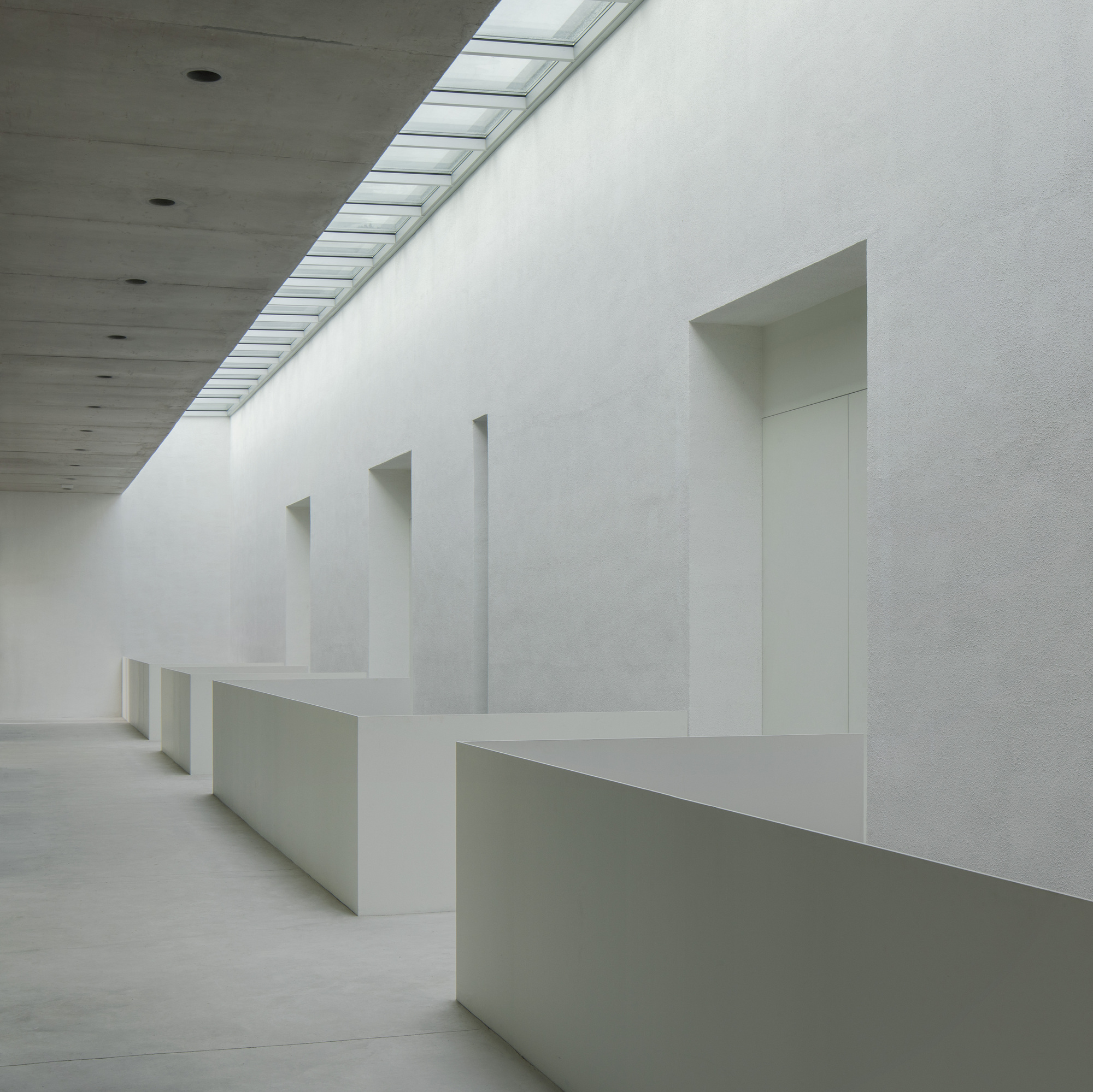 Interior hallway with recessed niches in white plaster walls beneath a linear skylight