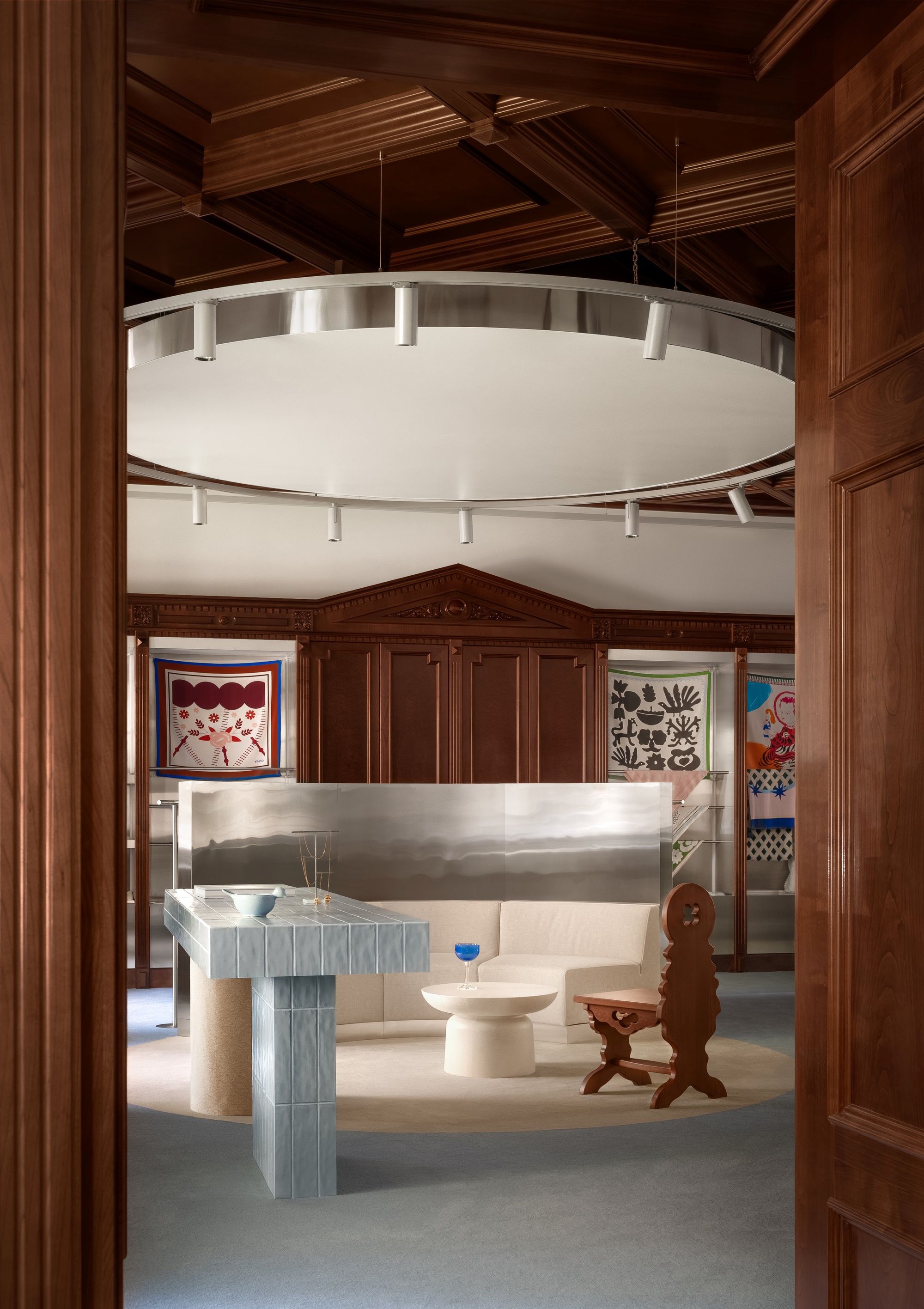 View through doorway to circular suspended lighting fixture above ribbed display tables and coffered ceiling