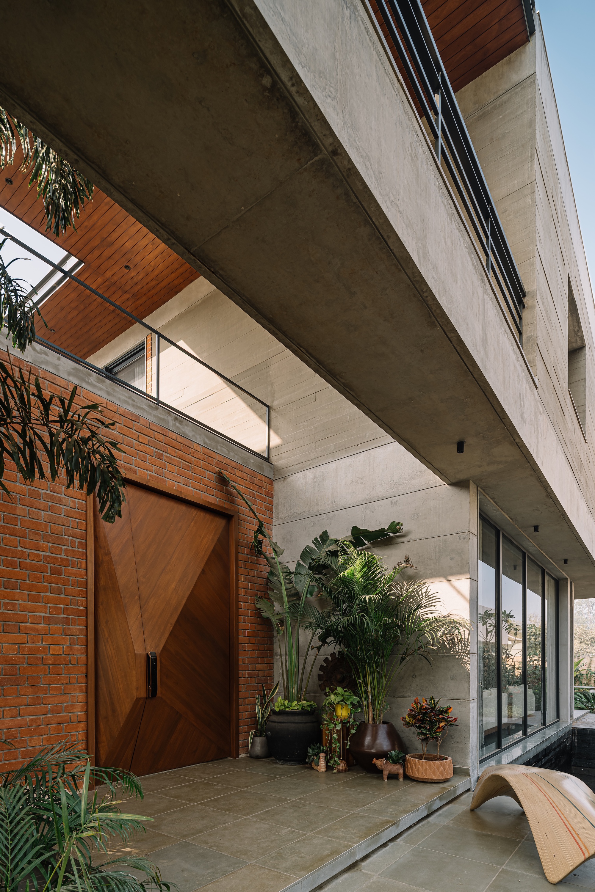 Covered entry courtyard with wooden pivot door, concrete soffit, potted plants and glazed upper volume