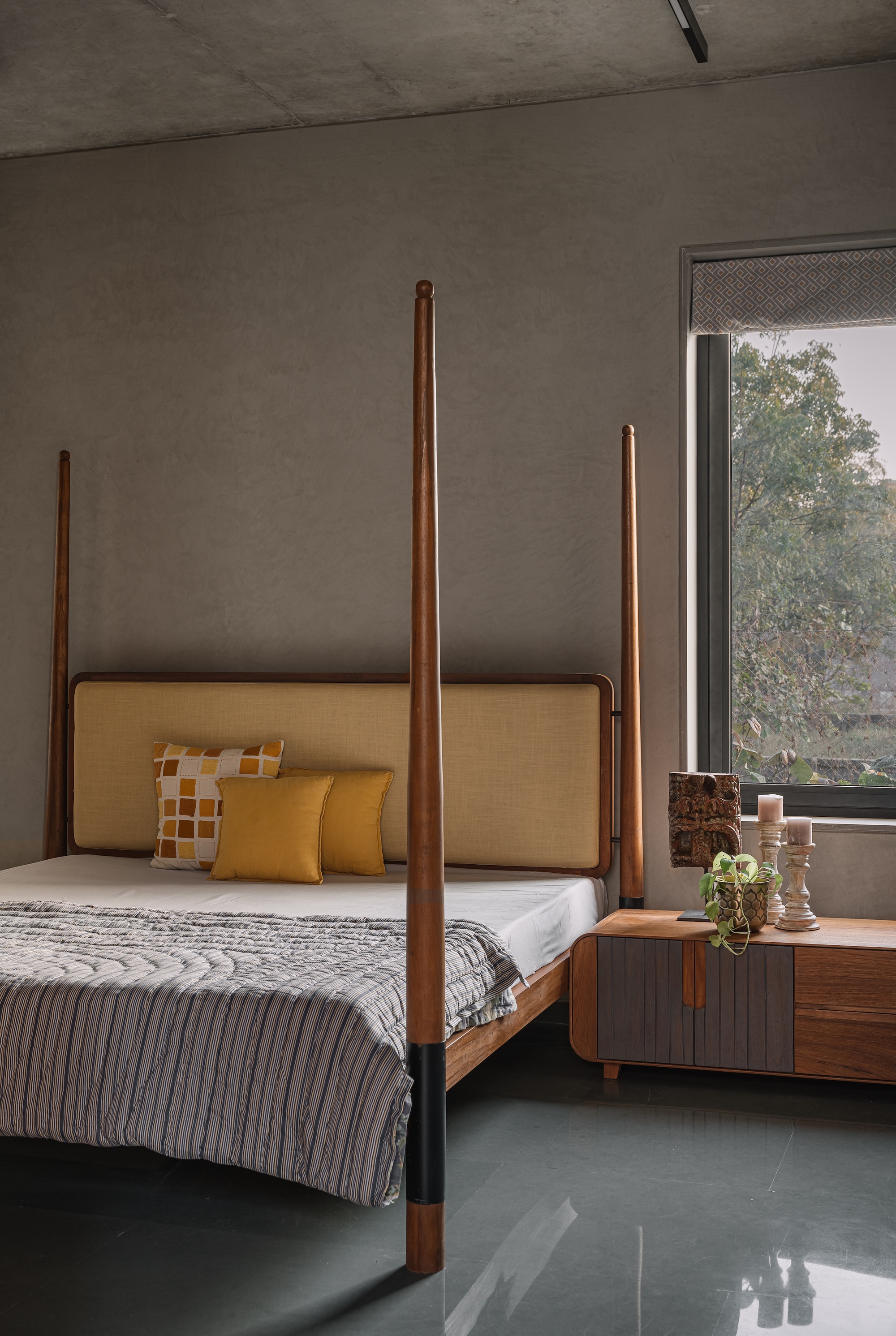 Four-poster bed with tall timber posts against a concrete wall with foliage visible through the window
