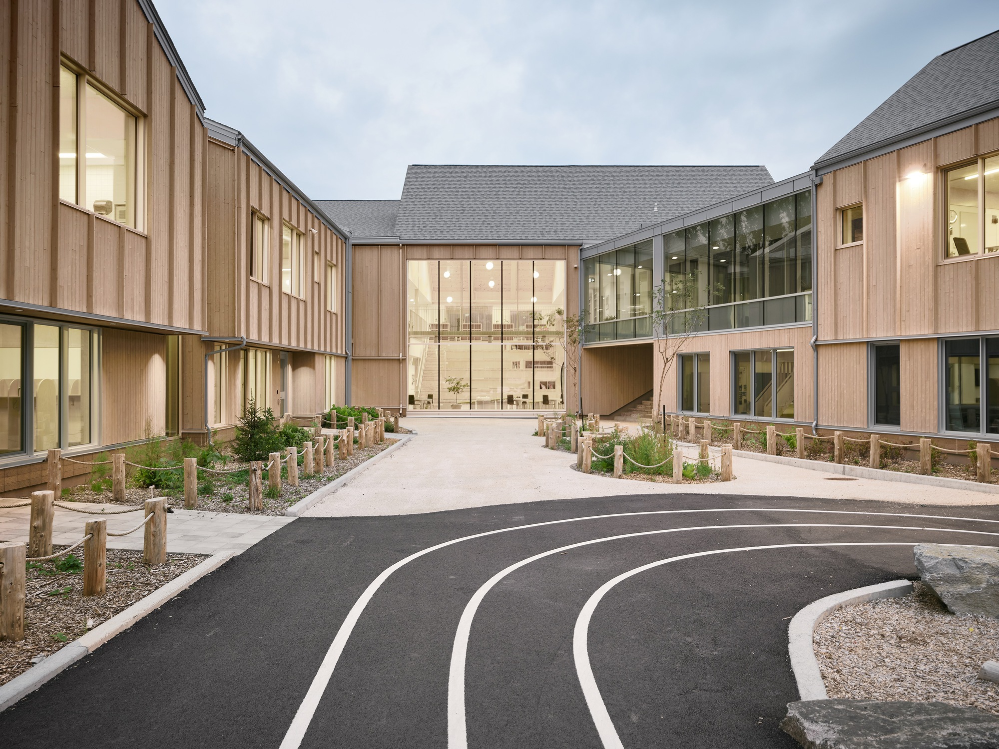 Central entrance with glazed facade and timber wings framing a curved asphalt path at twilight