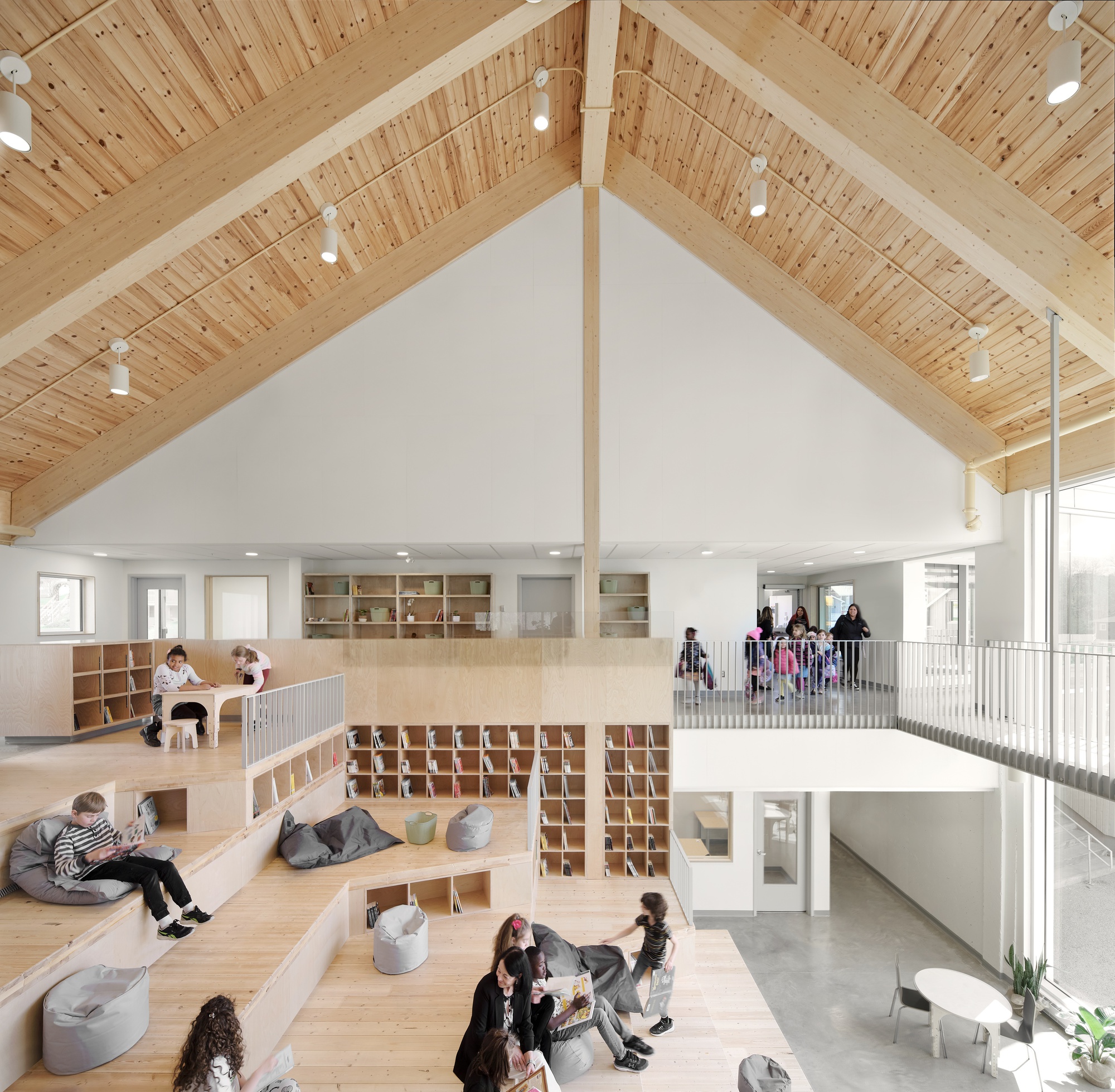 Double-height interior with exposed timber ceiling, plywood shelving, and children occupying tiered seating platforms