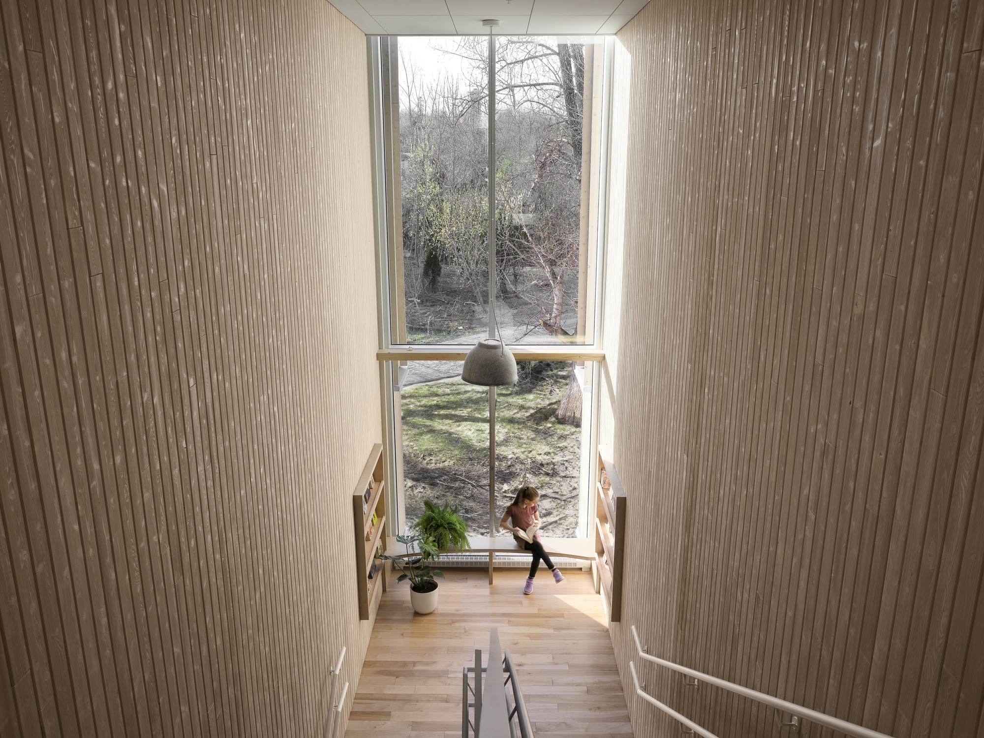 View down a timber staircase toward a tall window framing bare winter trees and a figure below