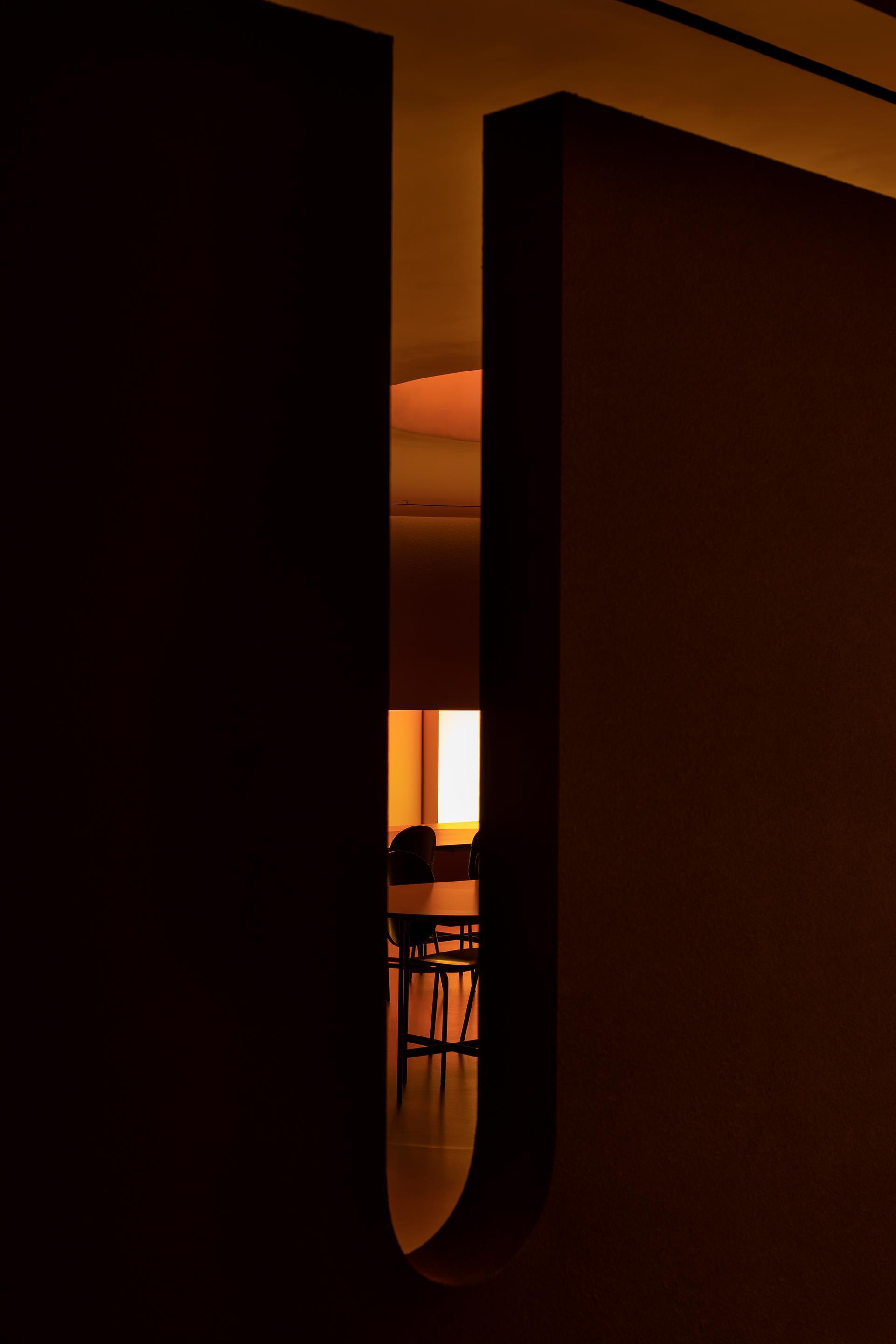 Narrow vertical opening framing a glimpsed interior with chairs under warm artificial light