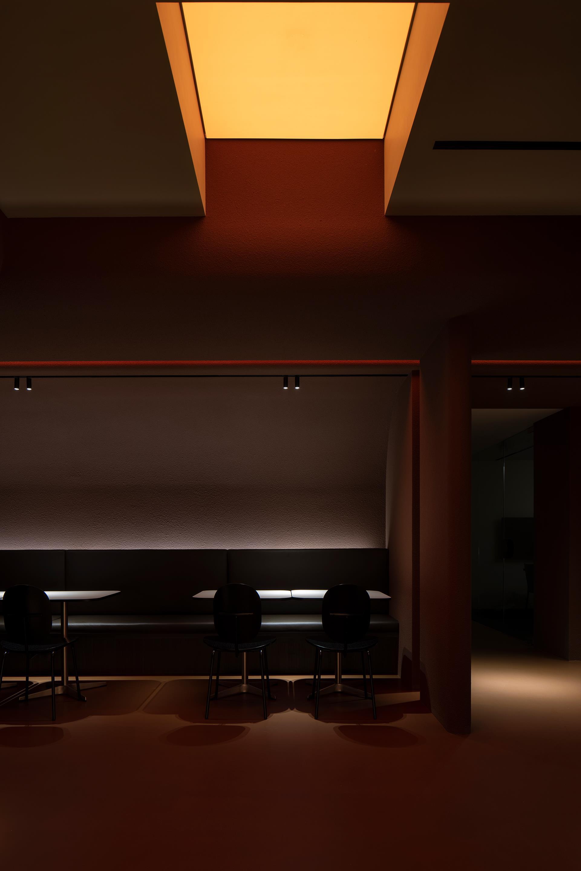 Dining area with long table beneath a recessed illuminated ceiling panel casting amber glow