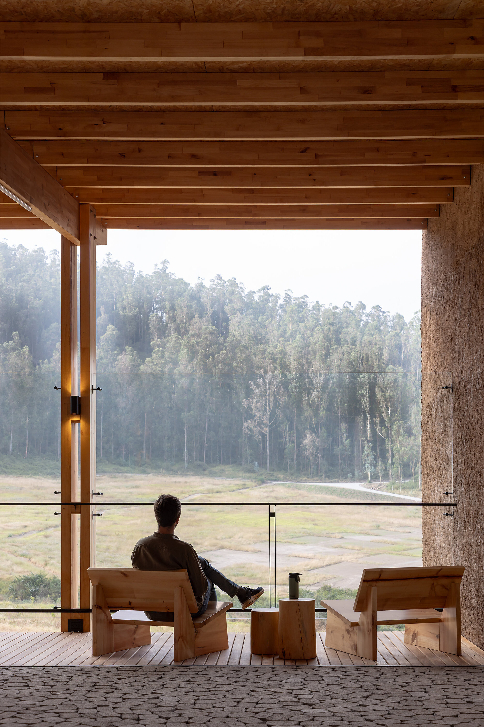 Covered veranda with exposed timber beams and person seated on wooden furniture overlooking misty fields
