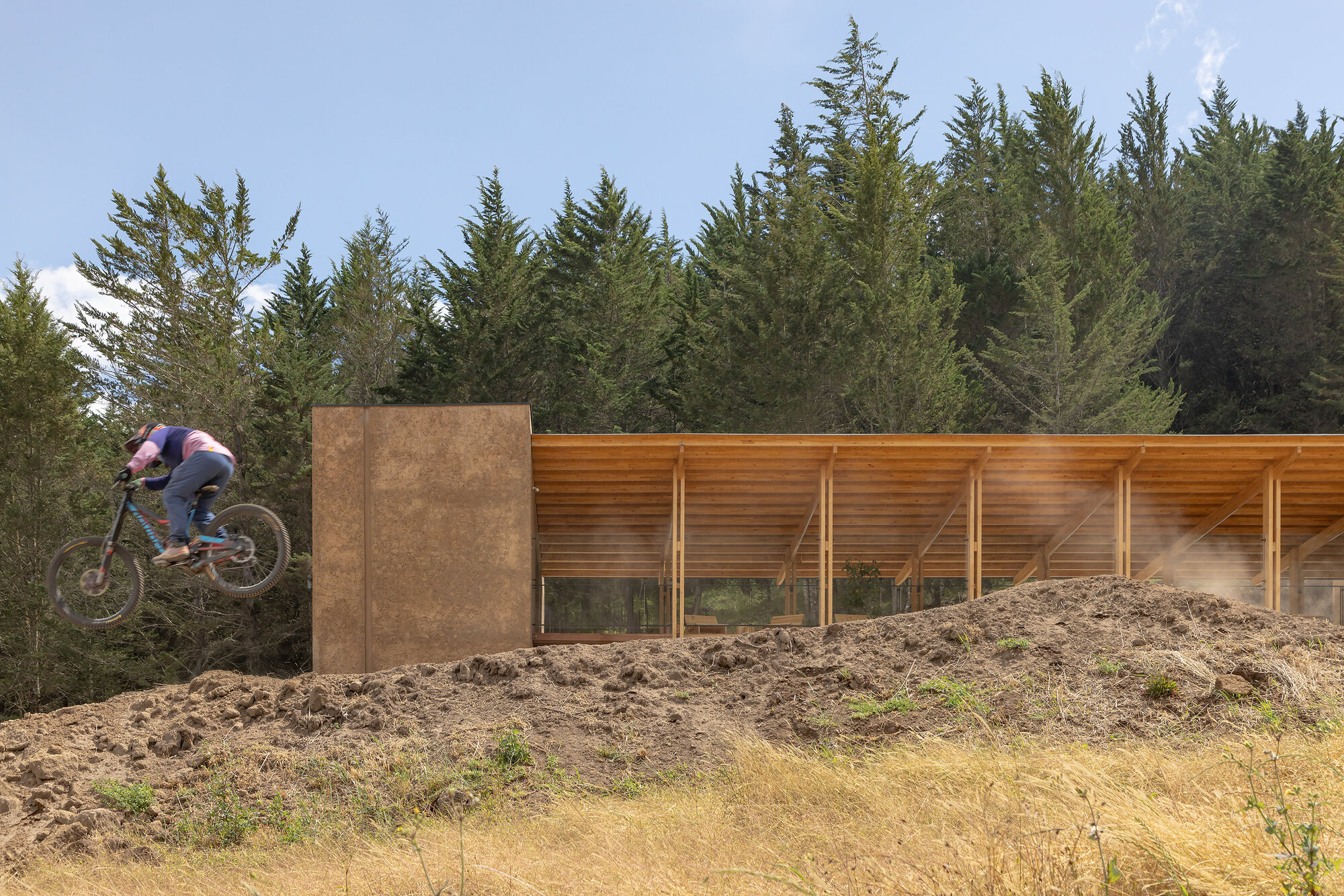 Cyclist jumping earthen berm beside rammed earth wall and timber-framed glazed facade in afternoon sun