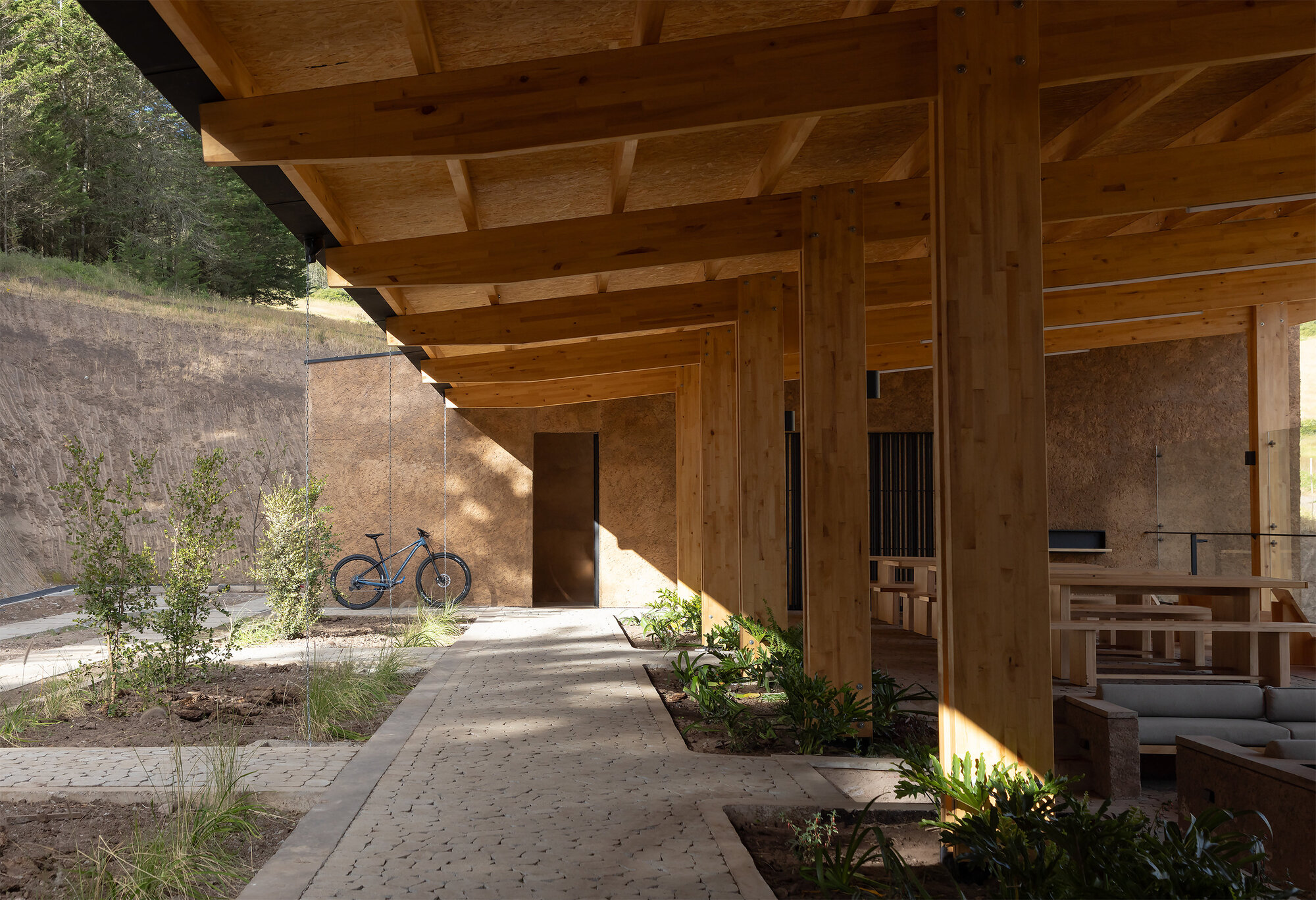 Covered walkway with timber beams and planted beds alongside a bicycle against a textured wall