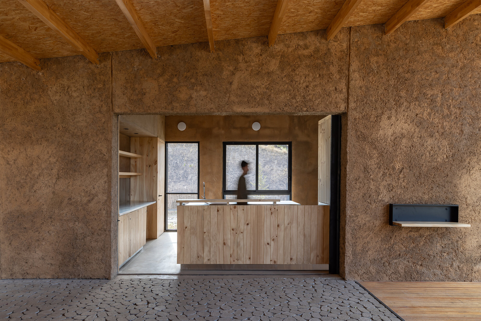 View from textured panel room through doorway to kitchen counter with person in motion inside
