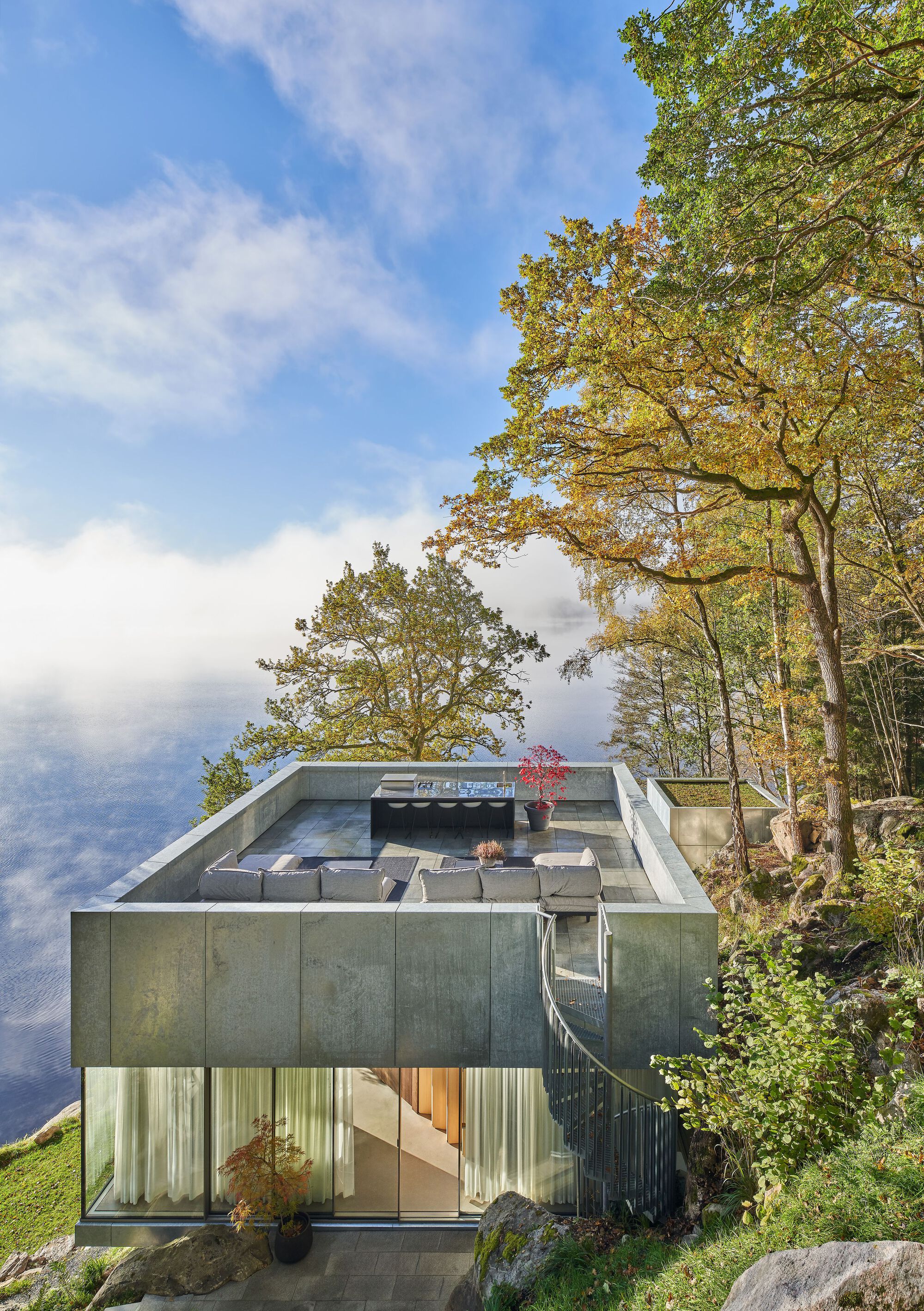 Two-story concrete volume with glazed lower level and rooftop terrace perched on a rocky slope among autumn trees