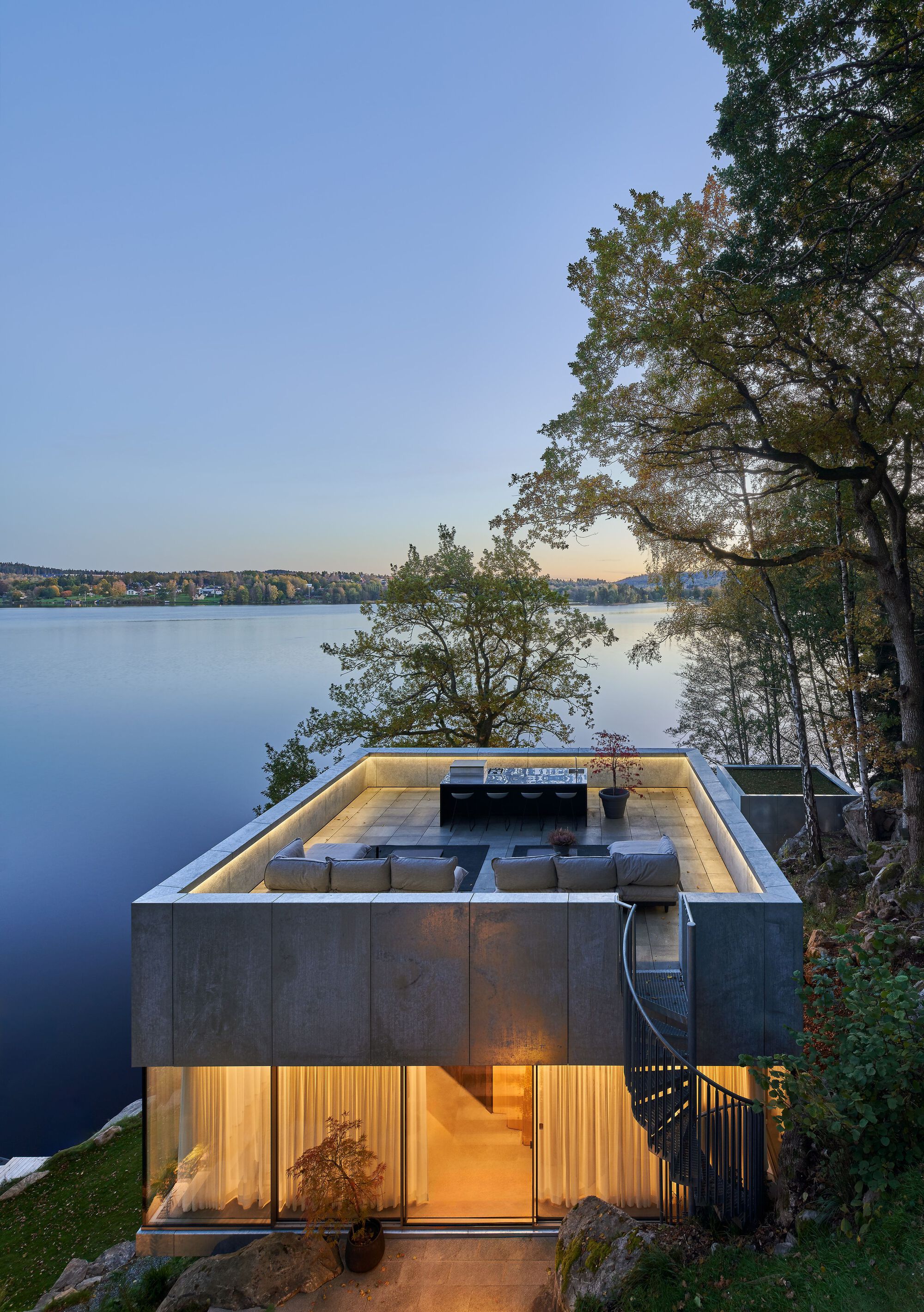 Lakeside facade at dusk with roof terrace above illuminated lower level timber and glass walls