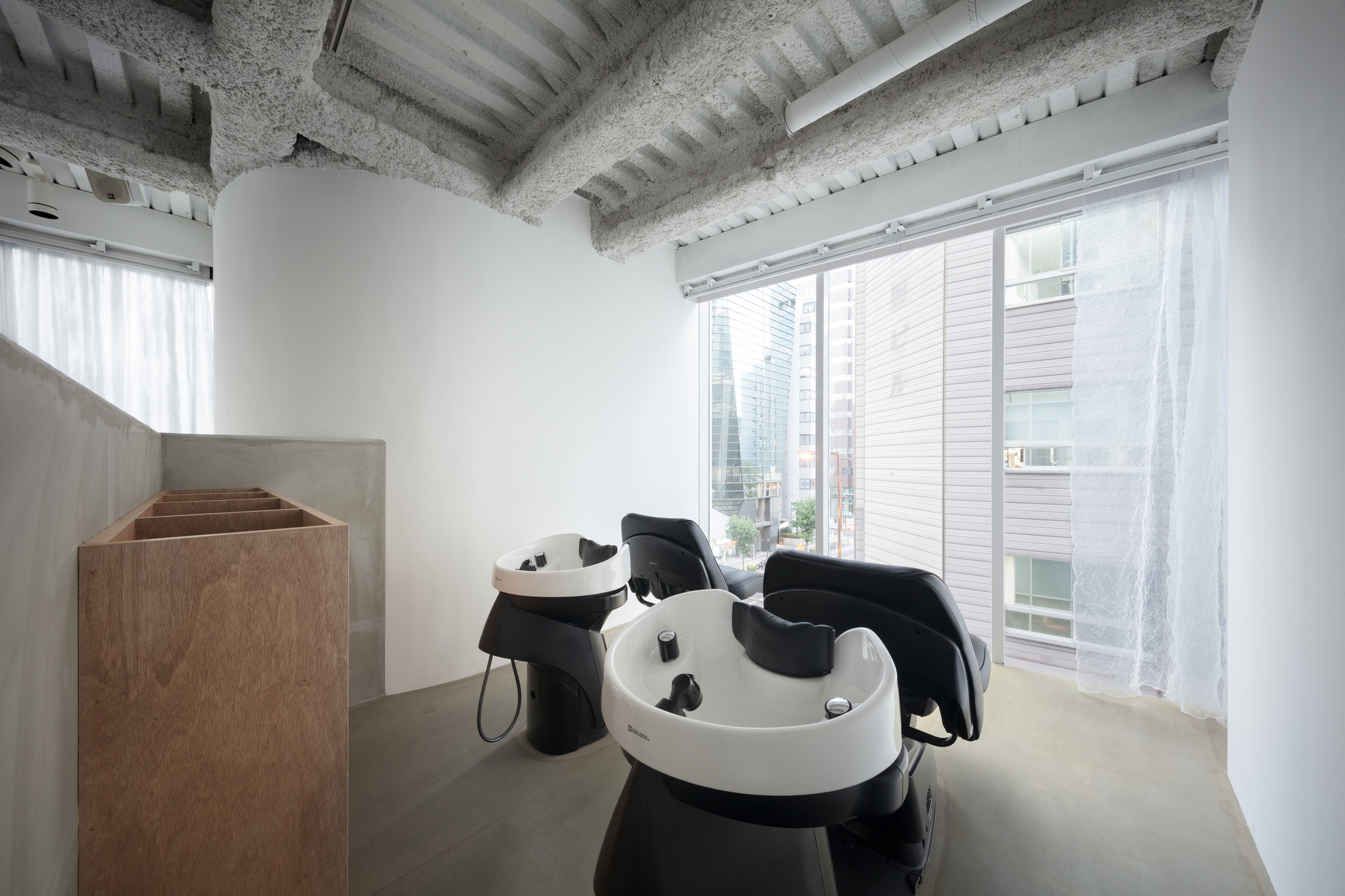 Hair washing stations with curved basins beneath exposed concrete ceiling beams and white curtains