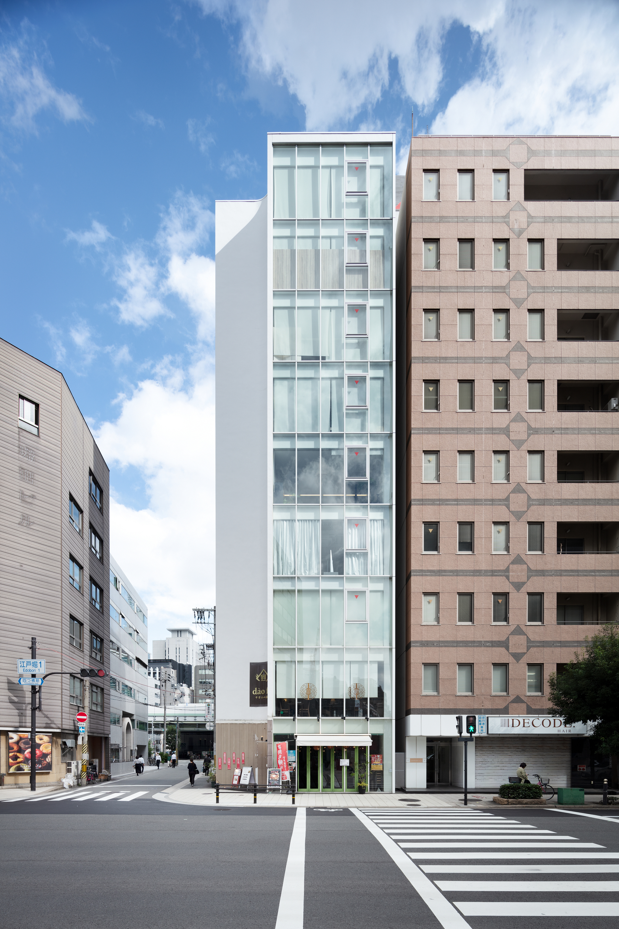 Narrow glass facade between neighboring buildings under a blue sky with scattered clouds