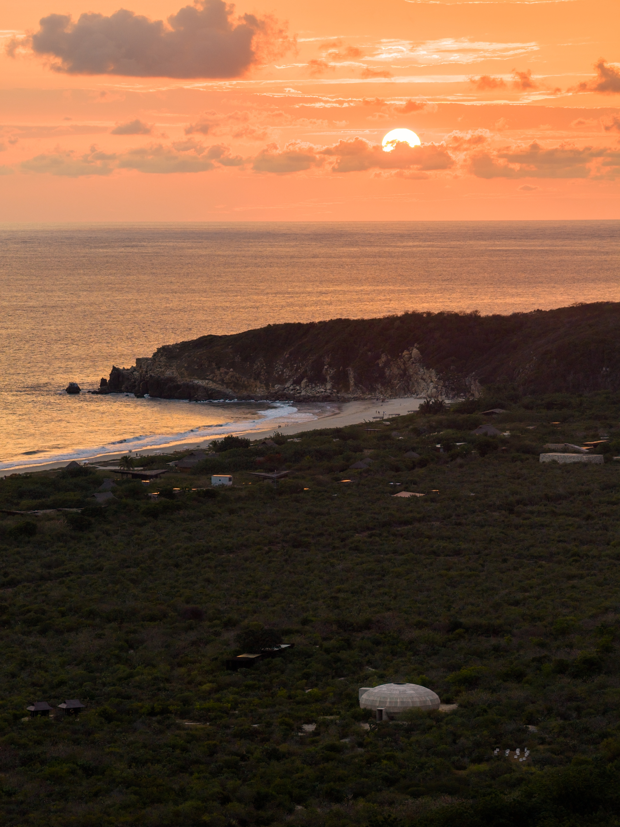 Aerial view of dome structure in dense vegetation with coastline and setting sun over water
