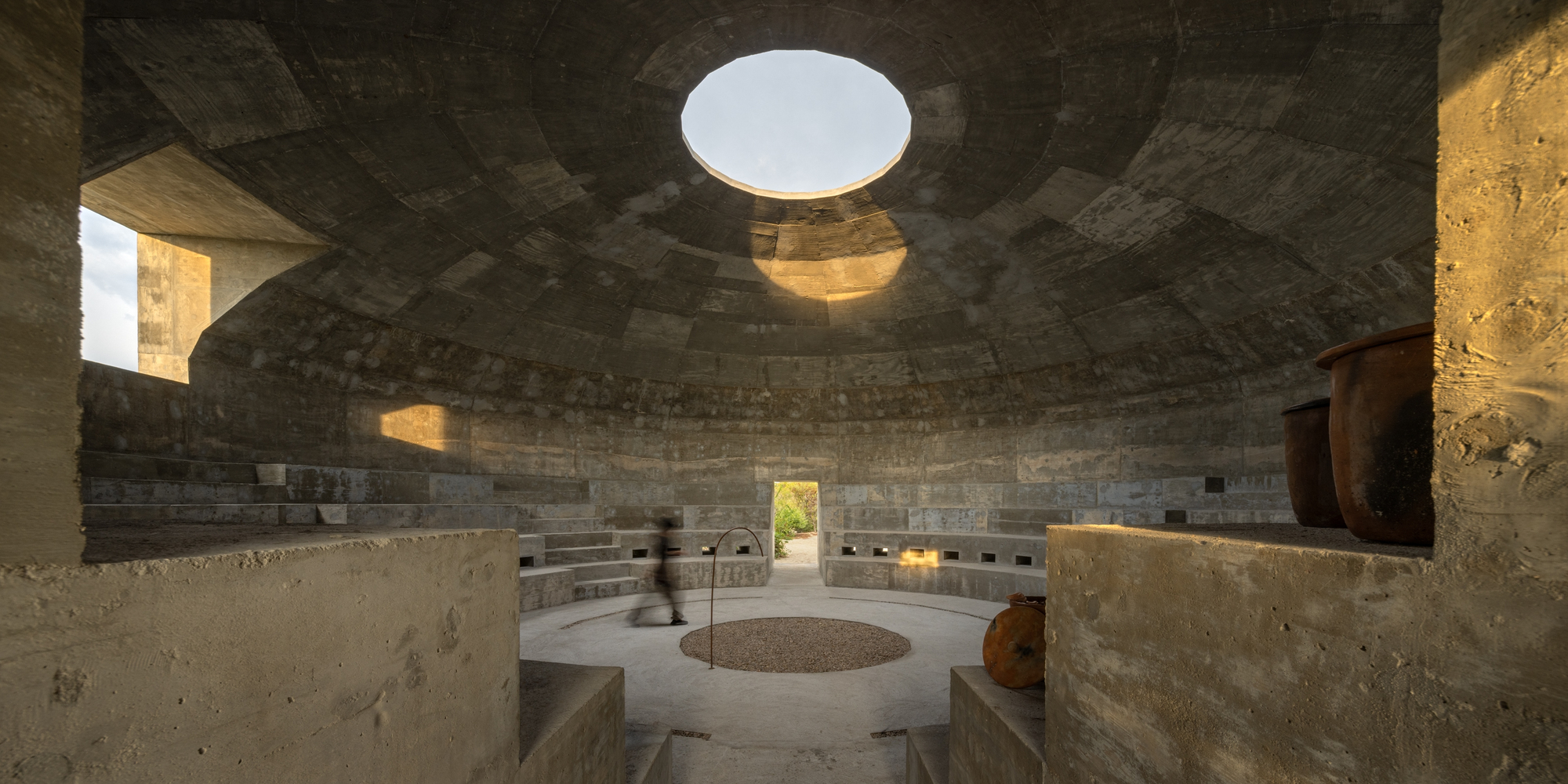 Interior of circular concrete chamber with tiered seating and central oculus casting afternoon light
