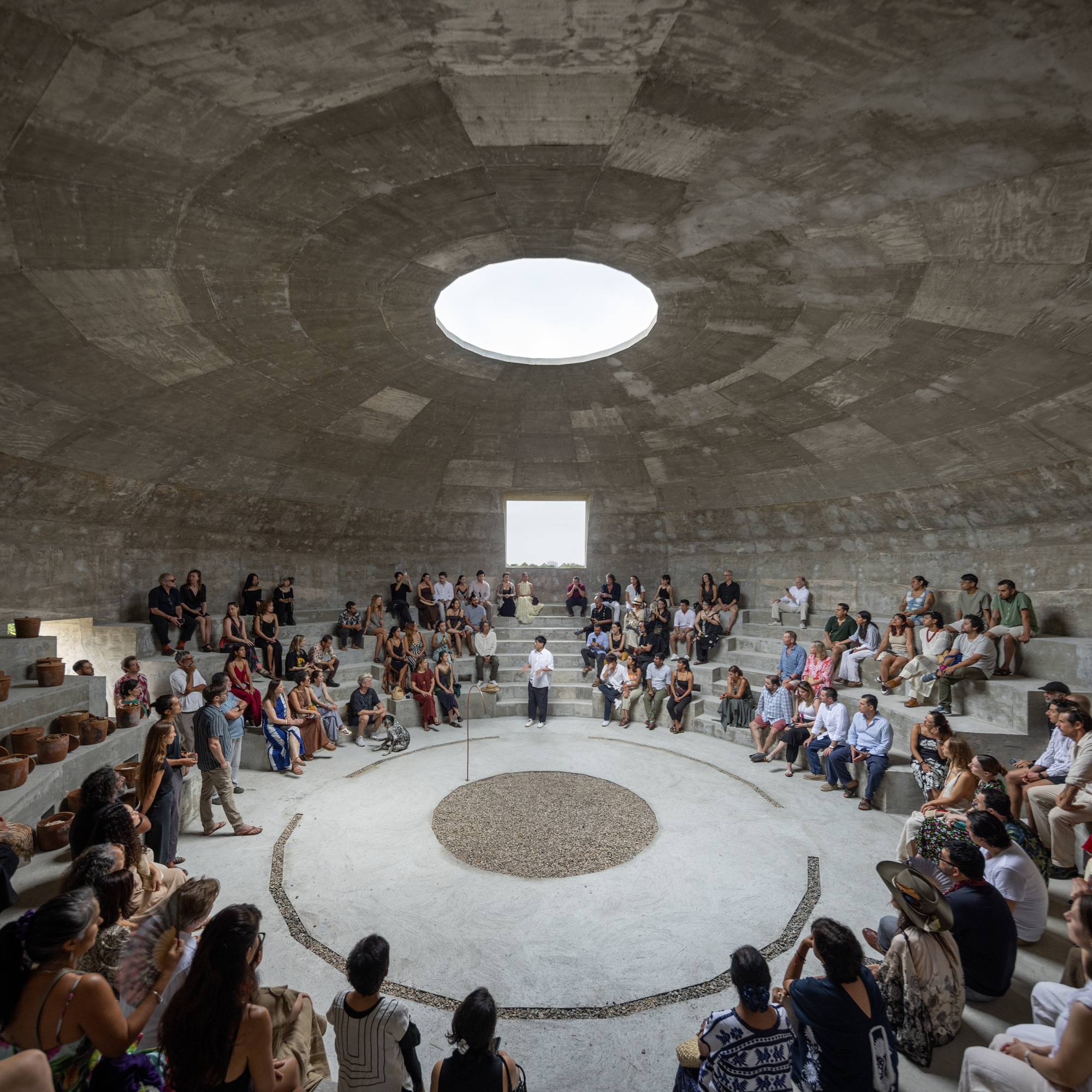 Amphitheater seating filled with visitors gathered around a gravel circle beneath the open oculus