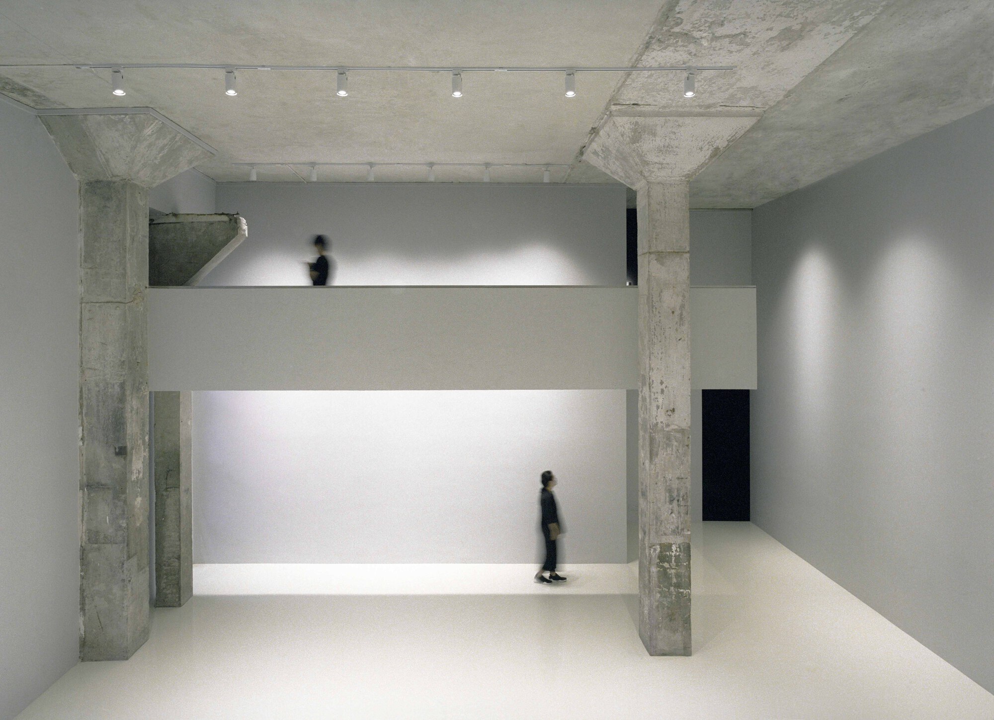 Double-height gallery space with illuminated horizontal display shelf and figure walking on upper level