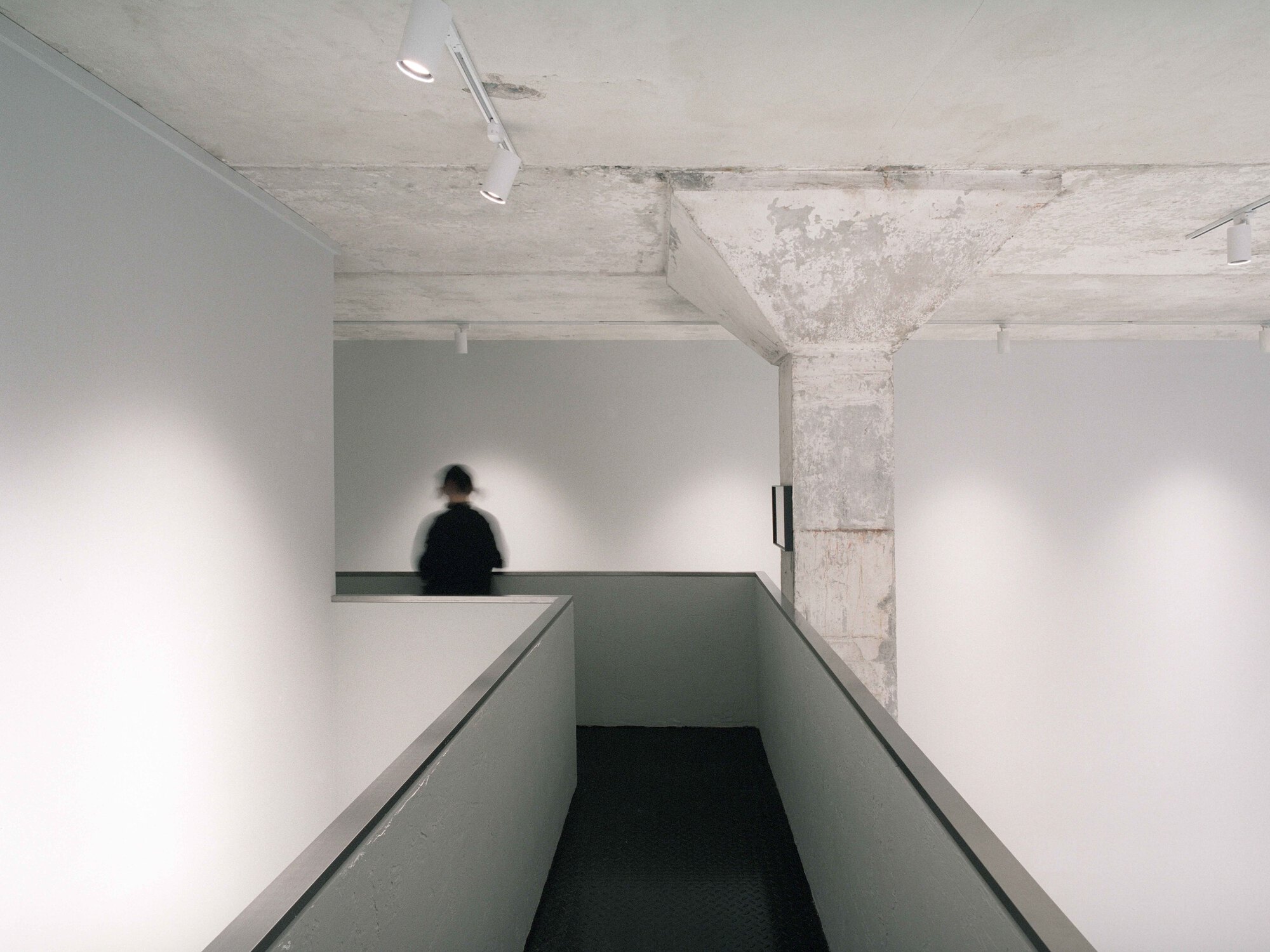 Ramp descending between white walls with concrete ceiling and figure visible on mezzanine beyond